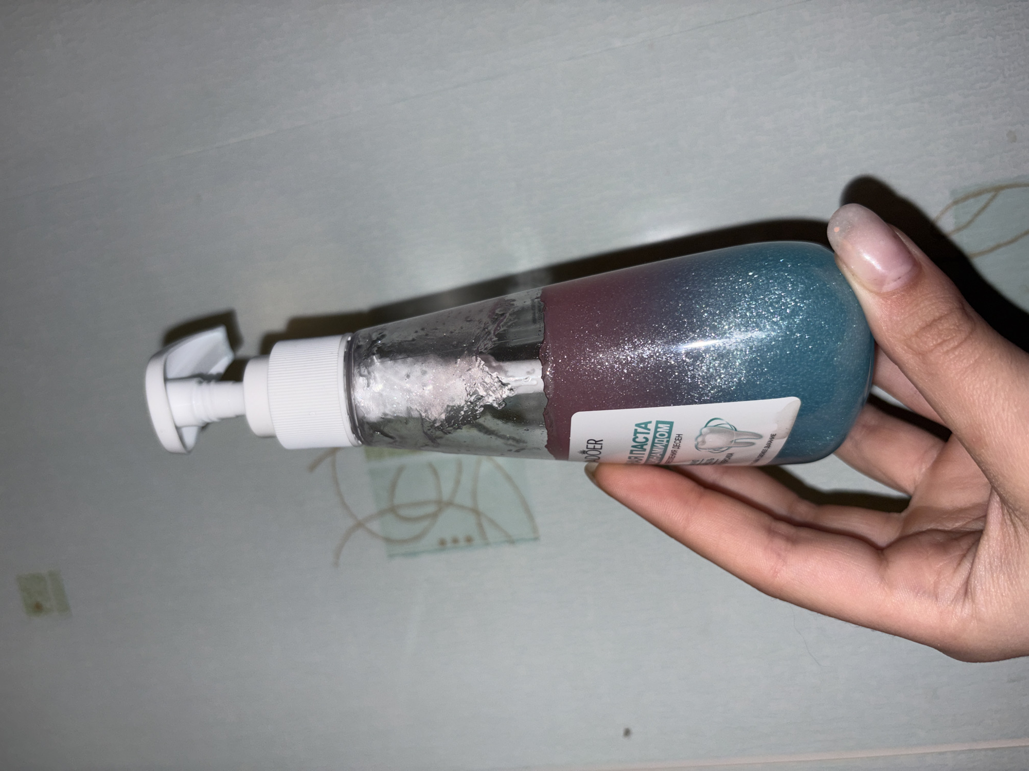A person's hand is holding a bottle of toothpaste. The toothpaste is multi-colored: clear on the top portion of the bottle, transitioning to purple, and then to a blue color on the bottom. The location may be Прикамский, Russia, but the environment is limited to the surface the bottle is resting on, which appears to be a white or light-colored countertop. The label on the toothpaste is partially visible and written in a language that appears to be Russian. The bottle has a pump dispenser on top. The person is holding the bottle horizontally in their hand, with the pump facing the left side of the image.