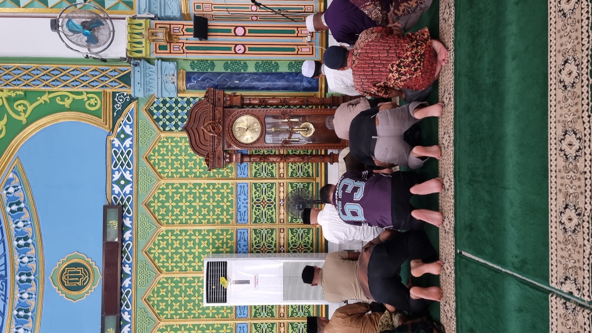 In the city of Keudekruenggeukueh, Indonesia, a group of seven men are performing the Islamic prayer known as "sujud" or prostration, their foreheads and noses touching a green carpet. They are positioned facing a large, ornate wooden grandfather clock. The men are dressed in various attire, including sarongs, shirts, and skullcaps. The interior of what appears to be a mosque features a vibrant and colorful wall adorned with intricate patterns in green, gold, and blue. An air conditioner unit stands to the left of the men, and a ceiling fan is visible on the right side of the frame. The overall environment suggests a religious gathering or service in progress.