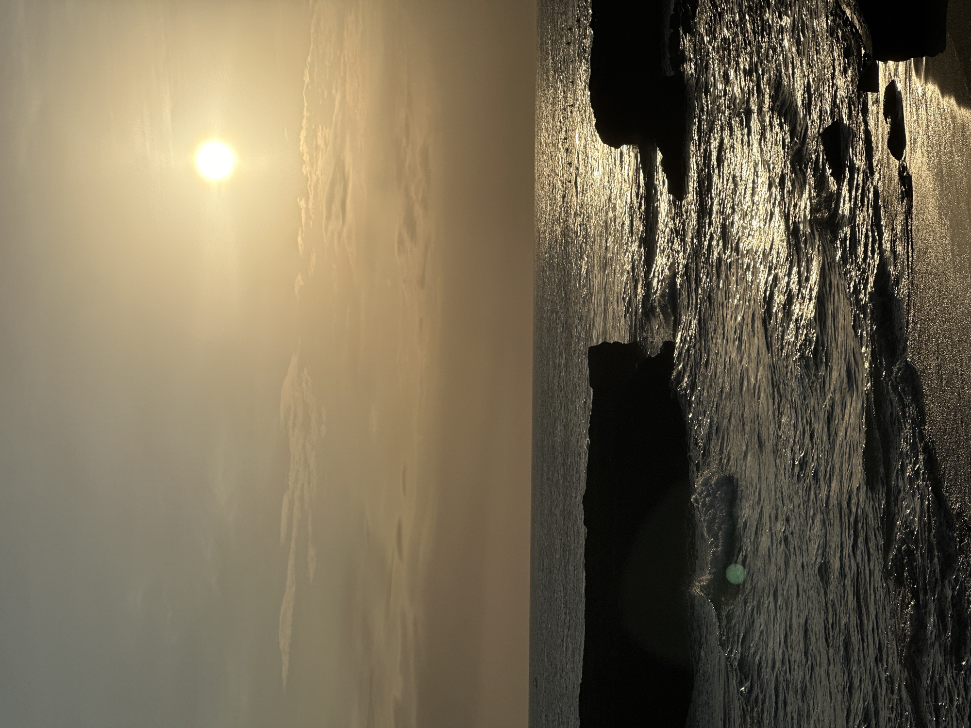 This vertical-oriented photograph captures a sunset over the ocean, likely at the beach. On the left side, a bright, hazy sun shines through a cloudy sky. On the right, the ocean surface reflects the sunlight, creating a shimmering, textured effect. Dark, rocky formations are present along the shoreline on the right. The image has a warm, golden hue due to the sunset. This photo was most likely taken in North Kuta, Indonesia.