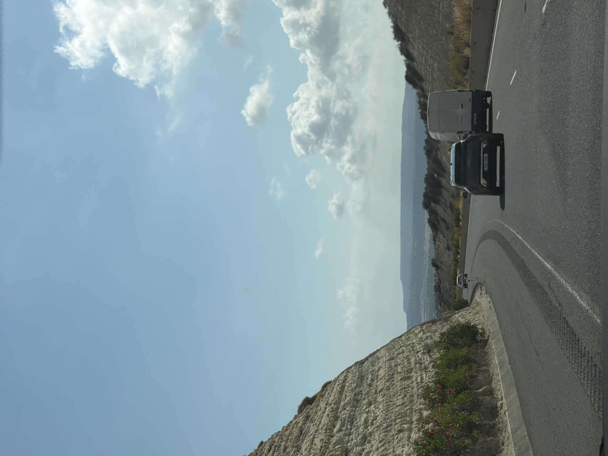 The image depicts a road scene with vehicles on a sunny day. A black SUV, with license plate TEF 560, is in the foreground, followed by a white van. The road curves to the right, bordered by a concrete barrier and a rocky, light-colored embankment on the left. Sparse pink-flowered bushes are visible along the base of the embankment. The background shows rolling hills with sparse vegetation under a blue sky with scattered white clouds. The location is Unknown, Unknown.