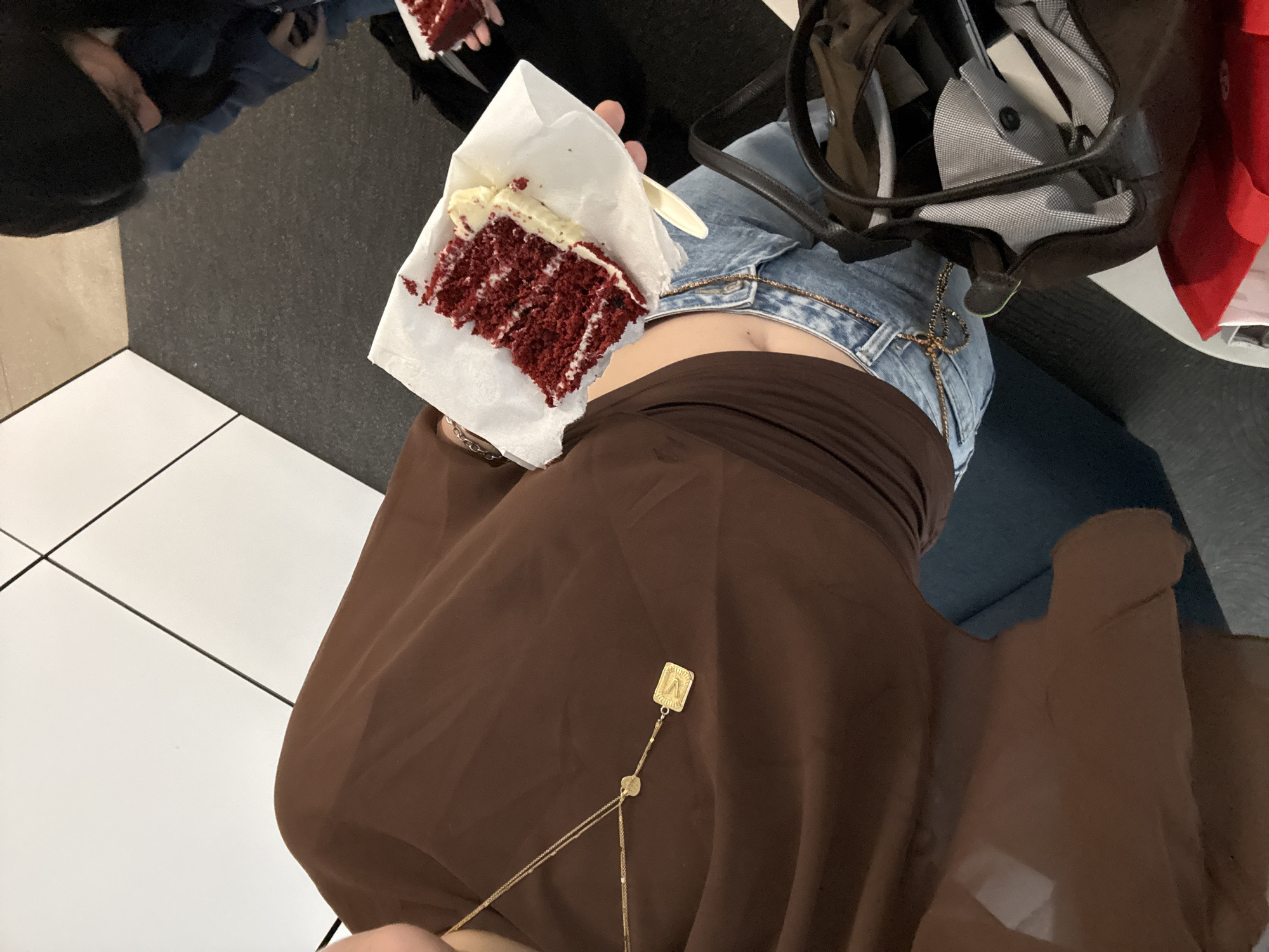 A person wearing blue jeans, a brown shirt, and a gold necklace sits, holding a piece of red velvet cake in a white paper wrapper. A black bag is next to the person on their right side. A second person is seated in the upper left of the image. The floor is made of white tiles and a dark gray surface. The image was taken indoors, likely in Mexico City, Mexico.
