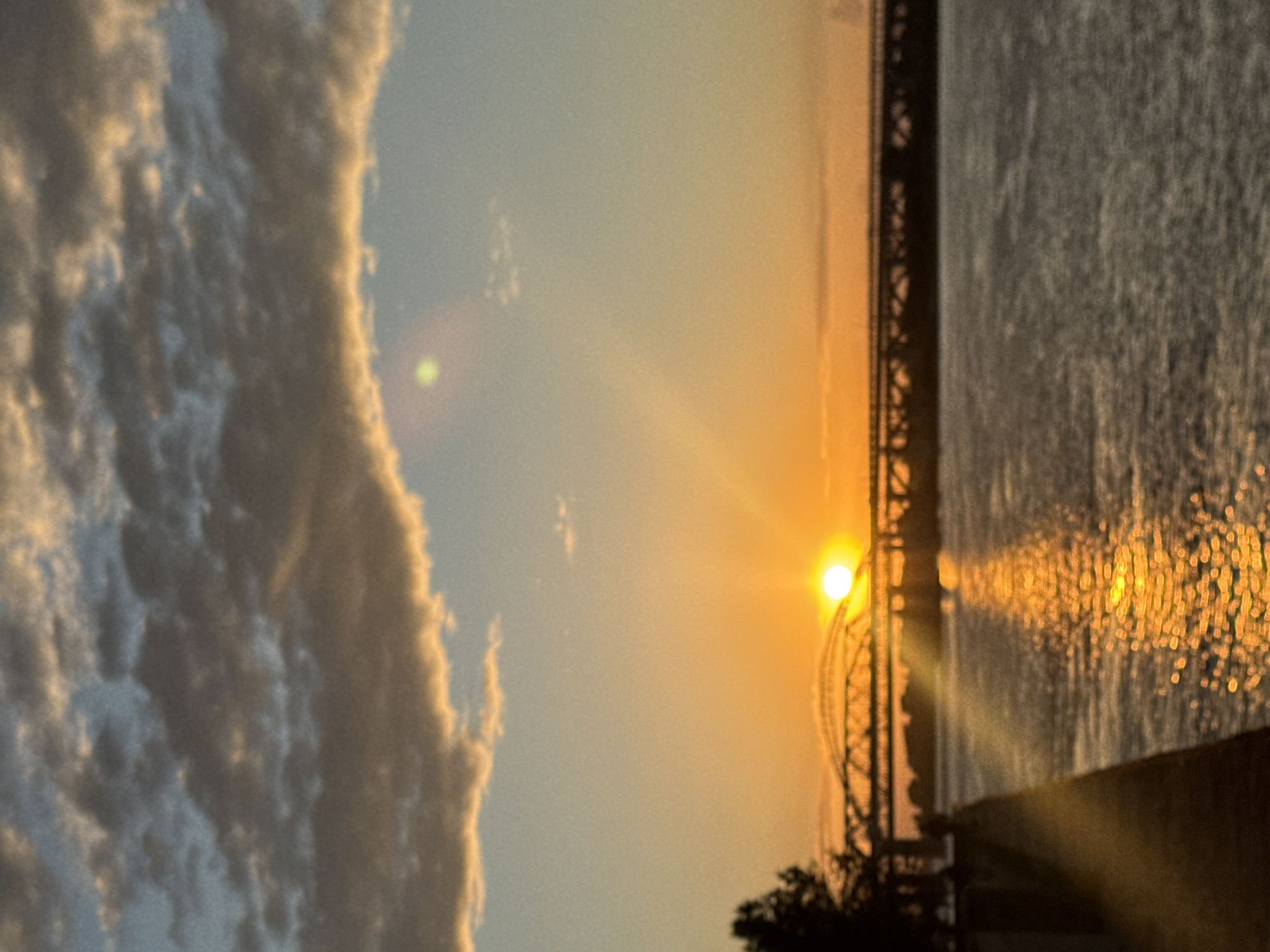The image depicts a sunset over a body of water, likely a river, with a bridge in the background. The sun is low on the horizon, casting a warm, golden glow across the scene. The water reflects the sunlight, creating a shimmering effect. Fluffy clouds dominate the left side of the frame, illuminated by the setting sun. The bridge appears to be a large, industrial structure with a distinctive arched section visible. The overall mood is serene and picturesque, suggesting a peaceful evening. The image is oriented vertically, implying it was taken with a phone in portrait mode. The lighting suggests it is either early morning or late evening. There is no visible text in the image. Given the location context of Nizhny Novgorod, Russia, this could be a view of the Volga River with one of its prominent bridges.