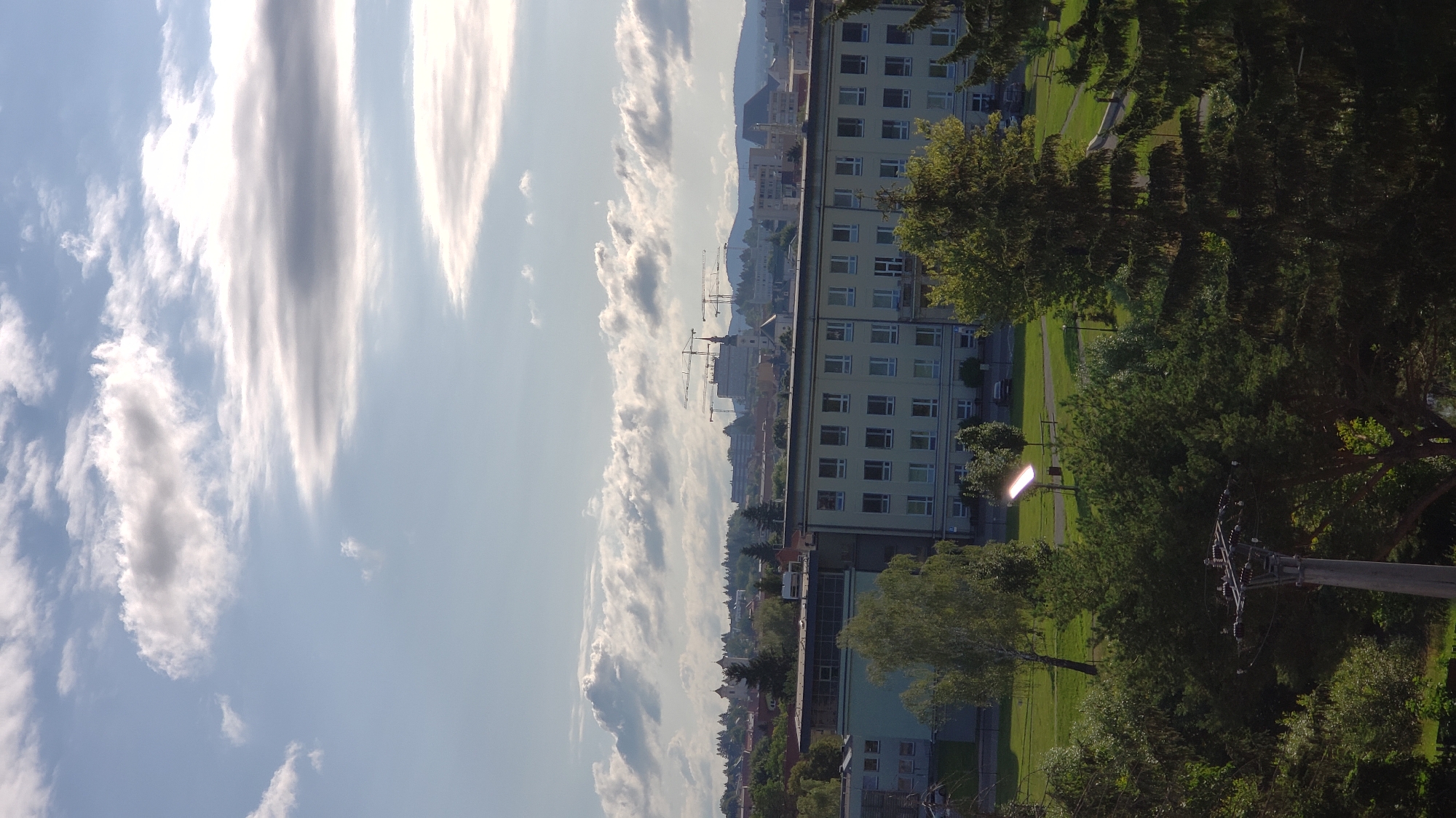 The image shows a view of a city from an elevated position, likely Prešov, Slovakia, on a partly cloudy day. In the foreground, a dense arrangement of evergreen trees dominates the lower portion of the frame. A single utility pole with wires and insulators is visible in the center-left foreground.
Beyond the trees, a light-colored, multi-story building with numerous windows is prominent. This building appears to be institutional or commercial, possibly a school or hospital, based on its scale and architecture. A well-maintained lawn surrounds the building, with a few scattered trees and a lamp post. In the middle ground, the cityscape unfolds, featuring a mix of buildings, including some with distinctive church spires. Construction cranes are visible in the distance, suggesting ongoing development. The background is characterized by rolling hills and mountains under a sky filled with a variety of clouds, ranging from wispy cirrus to larger, more dramatic formations. The overall impression is of a city viewed on a bright, possibly late afternoon, day.