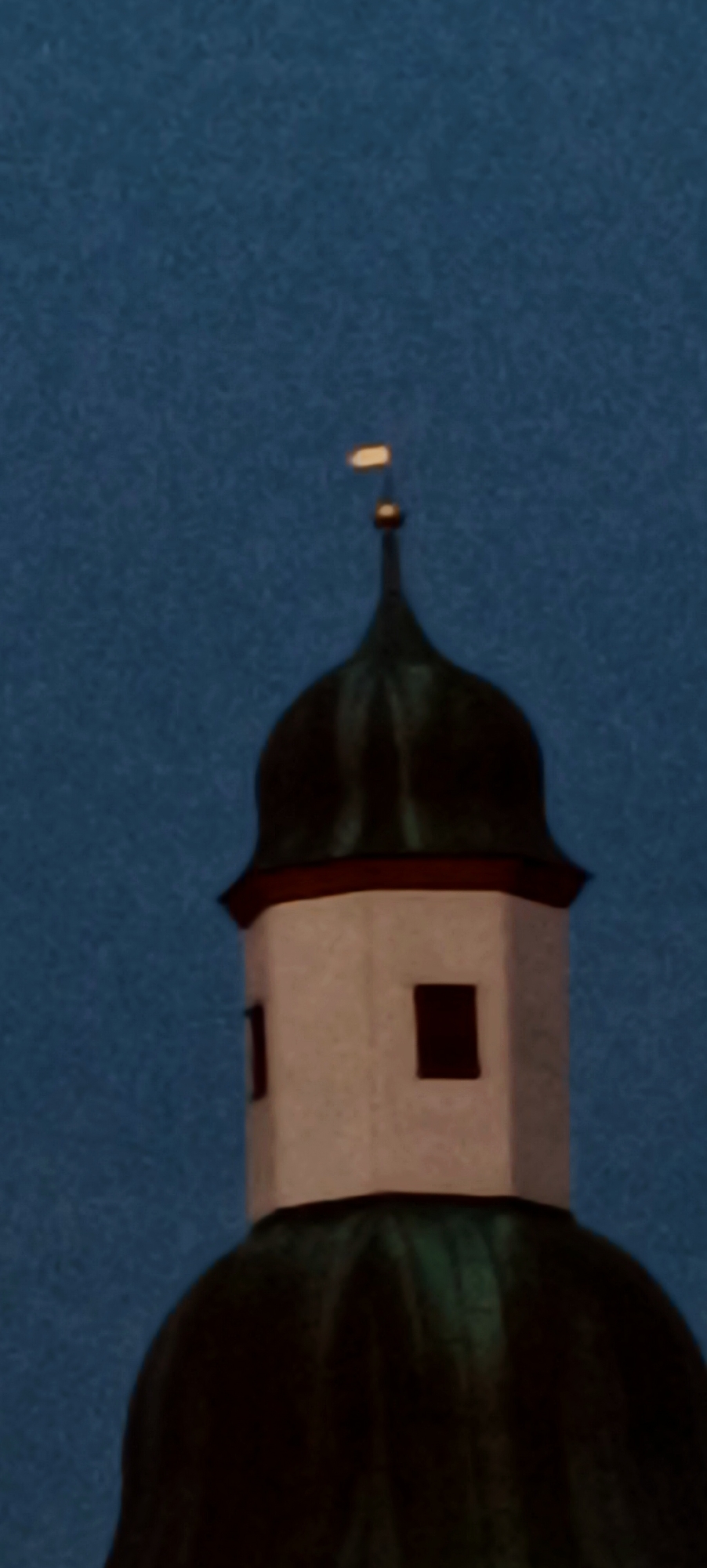 The image shows the upper part of a church steeple with a light blue, textured sky as the background. The steeple is composed of a white, polygonal base with dark rectangular windows on its visible sides. Above the white base, there is a dark green, rounded section, likely a dome or roof, topped by a slender dark spire. A small, yellowish, rectangular object is positioned at the very top of the spire, possibly a weather vane or a decorative element, illuminated from the left. The overall image is slightly grainy. This is the steeple of the Stadtkirche (City Church) in Winnenden, Germany.