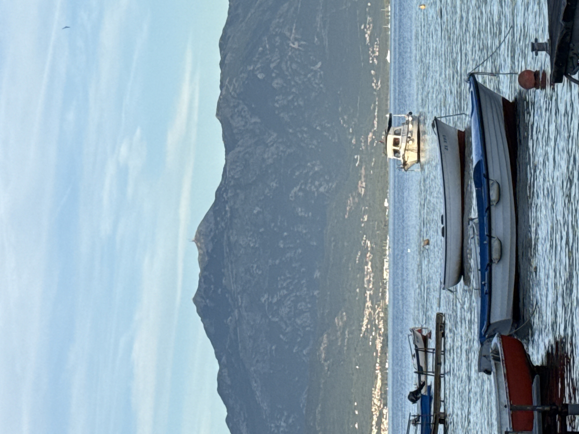 The image presents a wide view of a coastal scene in Herceg Novi, Montenegro, dominated by a large mountain range in the background, a town at its base, and multiple boats in the foreground on calm water under a partly cloudy sky.
The sky, occupying the top third of the frame, is a light blue with scattered, thin, horizontal stratiform clouds. A faint, dark linear trace, possibly a contrail, is visible in the upper right quadrant.
The background is dominated by a dark, rugged mountain range, primarily in shades of gray and dark blue, indicating rocky terrain and dense vegetation. The highest peak, slightly left of the image center, features a prominent summit. To its right, on a slightly lower peak, a vertical communication tower or antenna structure is visible.
At the base of the mountain, a coastal town is visible, characterized by clusters of light-colored buildings, some with red roofs, interspersed with green vegetation. The town stretches horizontally across the mid-ground, separating the mountain from the body of water.
The water, which makes up the lower half of the image, is calm with minor ripples, reflecting the sky. Its color ranges from deep blue in the distance to a lighter, greenish-blue in the foreground. Several white vessels, likely larger yachts or motorboats, are moored in the distance near the town.
In the immediate foreground, a collection of small boats is moored. A white cabin cruiser with a brown roof and a small mast is positioned in the mid-ground, floating freely. Closer to the viewer, two white rowboats are prominent. One on the left has a red hull bottom and a white topside, moored to a dark, wooden jetty structure partially visible in the lower left. Adjacent to it, slightly to the right, is another white rowboat with a blue trim and a dark (possibly red) hull bottom, secured by a rope extending towards the lower right. A red cylindrical mooring buoy floats near this boat. Further back and to the right of these, a third white rowboat with a dark hull bottom displays the registration number "ZL 197" on its bow. A yellow spherical buoy is visible floating further to the right. Portions of dark wooden pier structures are visible in the bottom left and bottom right corners, with a metal cleat present on the structure in the bottom right. No individuals are visible in the scene.