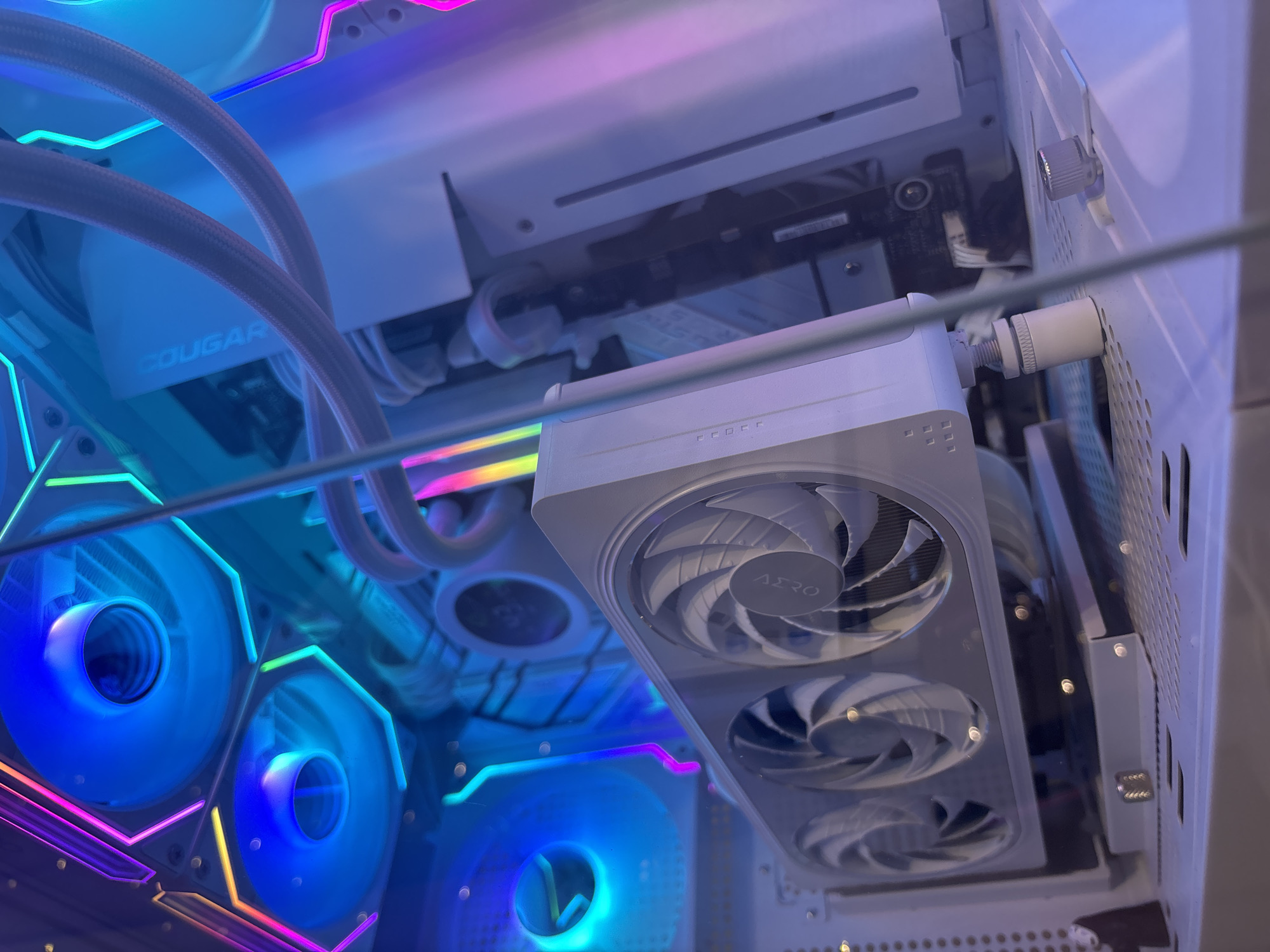 The image shows a close-up, interior view of a white computer tower with multiple fans and RGB lighting. A white graphics card with three fans is prominently displayed, angled towards the viewer. The fans on the graphics card are silver with a circular logo in the center. Above the graphics card, two large, round fans are visible, each with blue lighting and surrounded by hexagonal RGB lighting strips that cycle through pink, purple, and yellow hues. White ribbed tubes, likely for a liquid cooling system, are also visible. The interior of the computer case is white, with some black components and cables tucked away. The overall impression is one of a high-performance gaming or custom-built PC, with emphasis on aesthetics and cooling. The location is unknown.