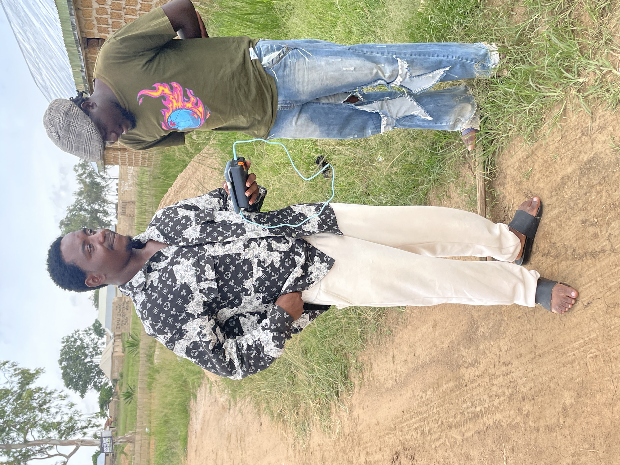 In the city of Durumi, Nigeria, two men stand in a dusty, outdoor environment, likely a village or rural area. The man on the left, with dark, curly hair, wears a black and white patterned shirt and light-colored pants. He holds a black and orange electronic device connected by a light blue wire to a similar device held by the man on the right. The man on the right, sporting a plaid flat cap, is dressed in a green t-shirt with a colorful graphic of a basketball engulfed in flames and distressed blue jeans. The background features a dirt path, overgrown grass, and simple brick buildings with metal roofs. Trees are visible in the distance, along with a sign on a tree to the left that reads "GOD IS NOT A MAN". The scene appears to be a moment of technological interaction or demonstration in a communal setting.