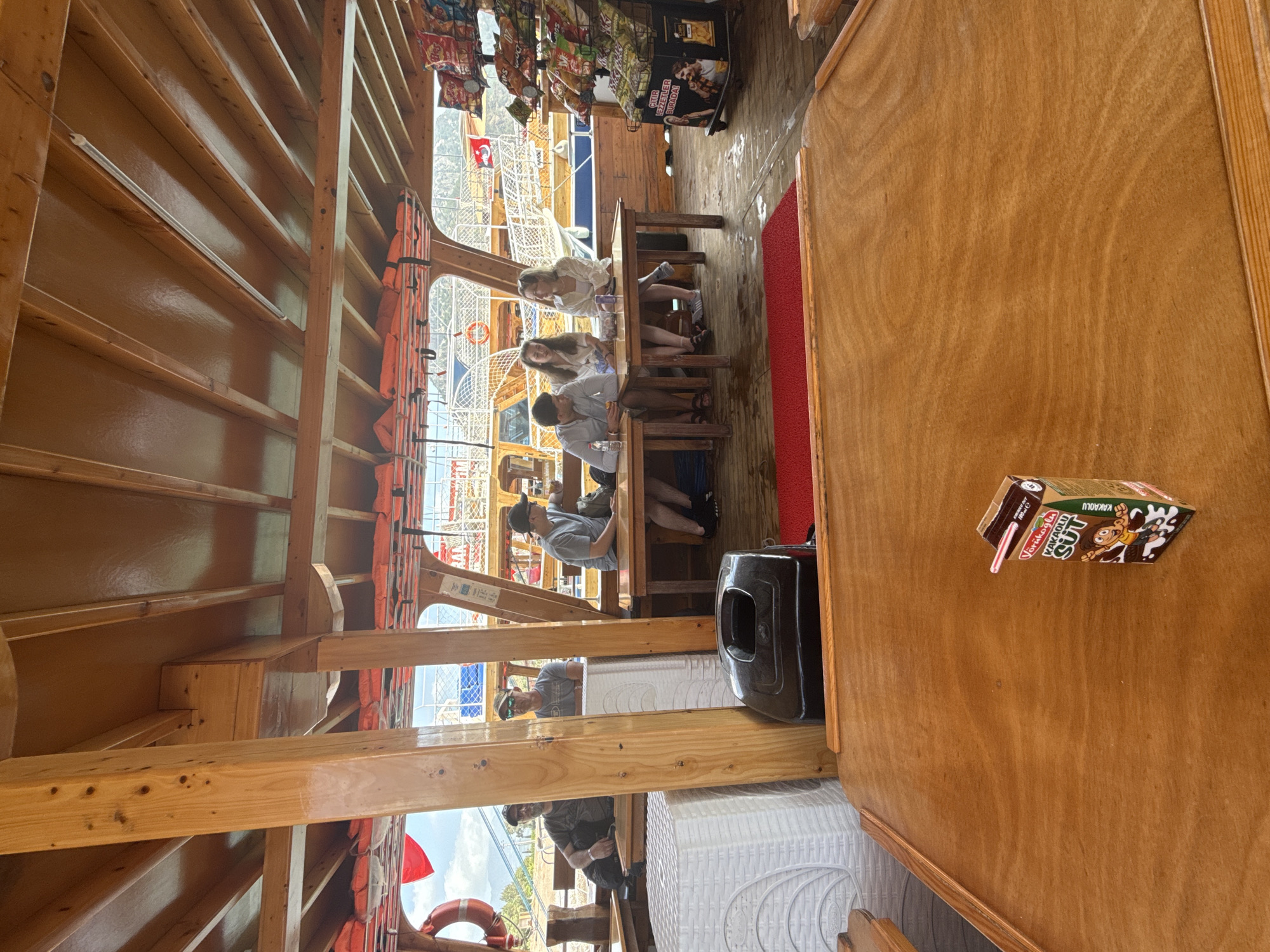 The image depicts the interior of a boat in Kumluca, Turkey. Passengers are seated at tables in a wooden structure. Life jackets hang on the side. A drink box rests on a wooden table.