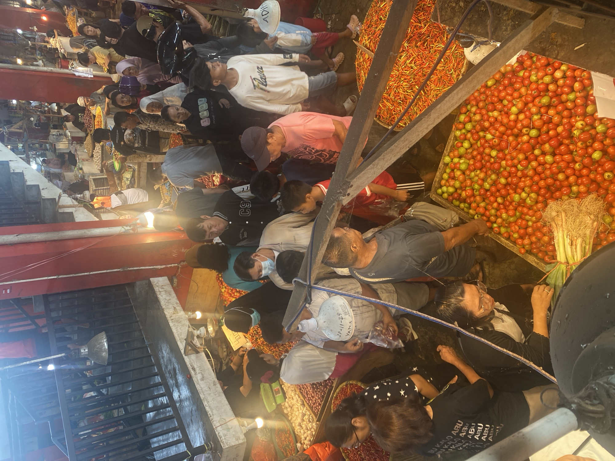 This image depicts a bustling indoor market scene, likely in Manado, Indonesia, given the context. The market is filled with vendors and shoppers navigating narrow aisles and crowded spaces. Various produce, primarily chilies and tomatoes, are prominently displayed in large baskets and crates. People of diverse ages and backgrounds are seen interacting, examining goods, and making purchases. The atmosphere appears lively and energetic, with artificial lighting illuminating the stalls. The architecture suggests a permanent market structure with metal railings and red-painted pillars. Some individuals are wearing masks, indicating a possible ongoing public health concern.