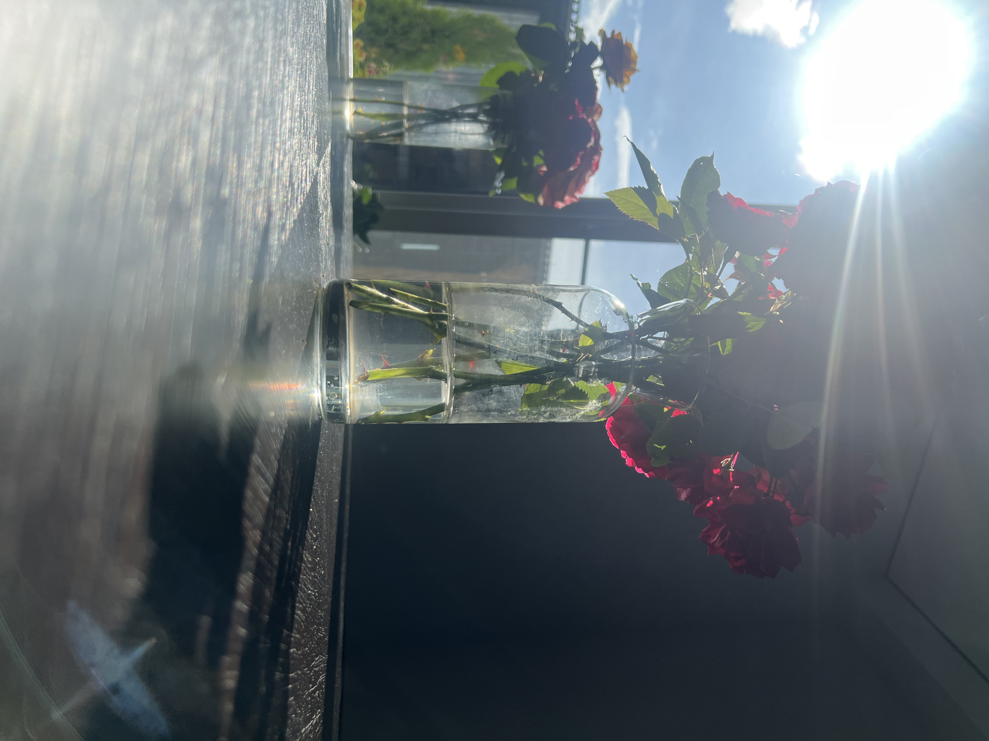 A close-up shot captures a glass jar containing a few rose stems with thorns and leaves submerged in water. The jar itself is partially filled, with the water reflecting light. In the background, another glass jar with a bouquet of reddish-orange roses is visible, also positioned indoors, likely near a window. The overall scene is illuminated by bright sunlight streaming in from the right, creating lens flare and casting shadows. The sky appears blue with scattered clouds. The image conveys a sense of natural beauty and a peaceful, indoor setting, possibly a home or studio in Minzag, Russia. The strong sunlight suggests it is daytime, and the bright conditions imply fair weather. There is no visible text in the image.