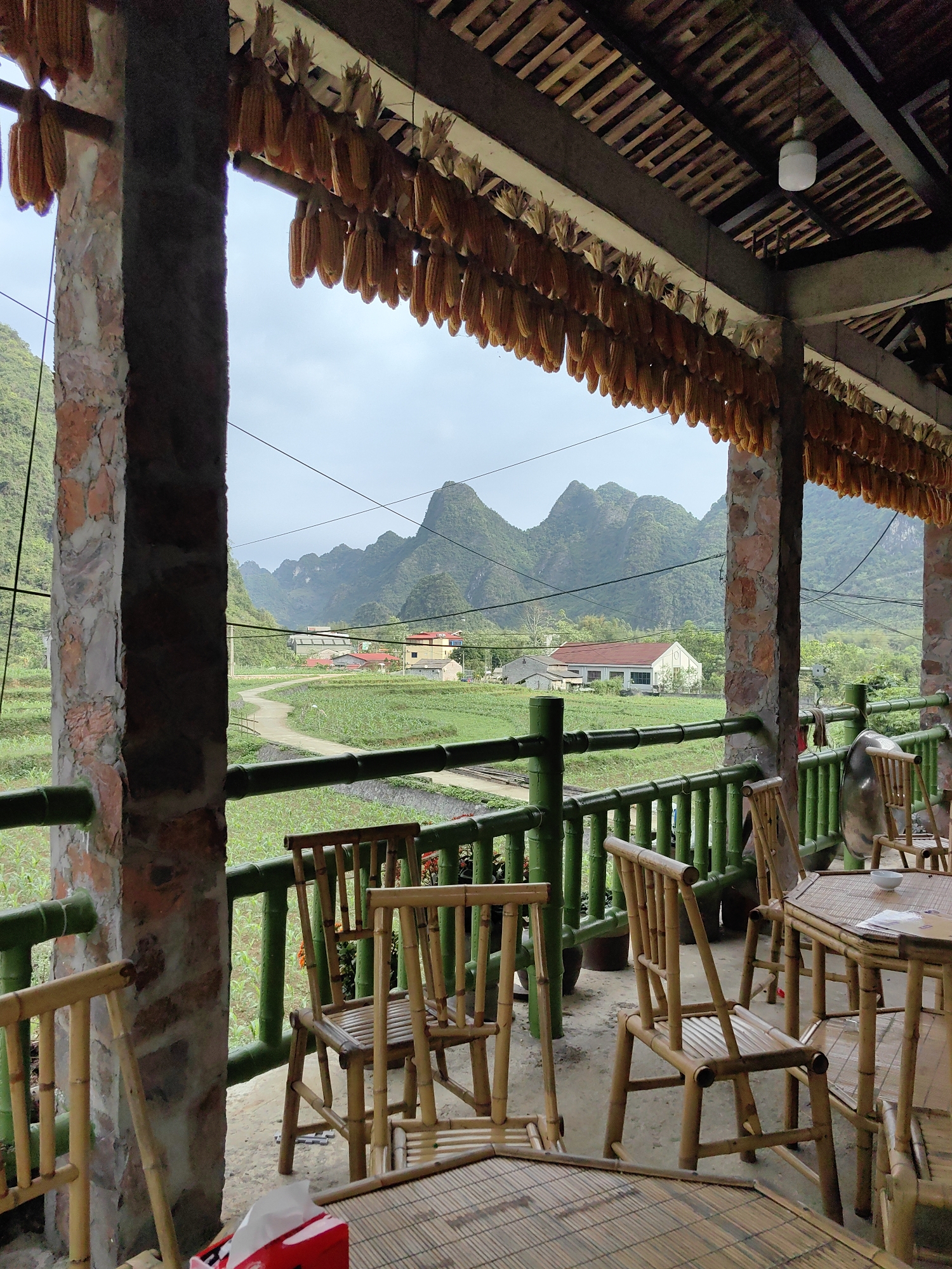The image shows an outdoor dining area with bamboo furniture, likely a restaurant or cafe, in Xã Đàm Thủy, Vietnam. Corn cobs hang from the ceiling. A view of mountains, fields, and rural buildings is visible through the open space. The tables are set with simple place settings. The furniture and railing are mostly green and brown. The sky is overcast.