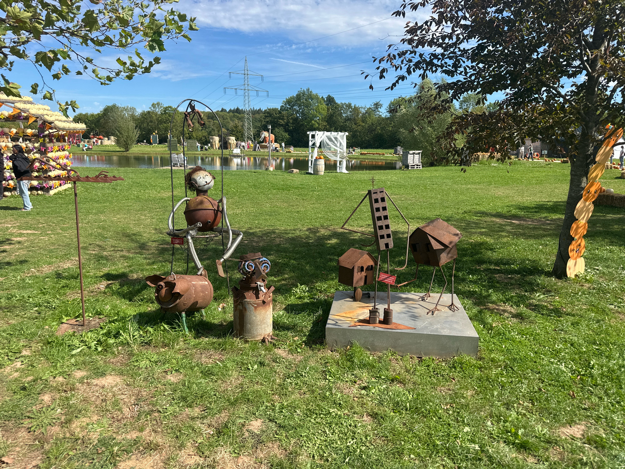 The image depicts an outdoor autumnal scene, likely part of a seasonal attraction, possibly a pumpkin festival, in the city of Mechernich, Germany. In the foreground, to the left, is a whimsical metal sculpture of a pig, seemingly made from repurposed materials like an old oil drum. Next to it stands another sculpture of a figure, also made of metal, with a round head and a torso resembling a pot, sitting on what appears to be a swing. To the right, a metal sculpture depicts a family of houses with spindly legs, standing on a grey platform. Beyond these foreground elements, a body of water, possibly a lake or pond, is visible. In the middle ground, various decorations and structures can be seen, including hay bales, wooden crates, and a white archway draped with fabric, suggesting a wedding or event setup. Further in the background, trees form a dense line, and power pylons are visible against a blue sky with scattered clouds. Several people are scattered throughout the scene, some walking or standing near the pond and decorations. On the far left, a display of pumpkins and gourds is arranged, with a person in the foreground looking at them. The ground is covered in green grass, with patches of exposed soil and scattered weeds. The overall impression is a festive, outdoors event with a focus on seasonal decorations and creative art installations.