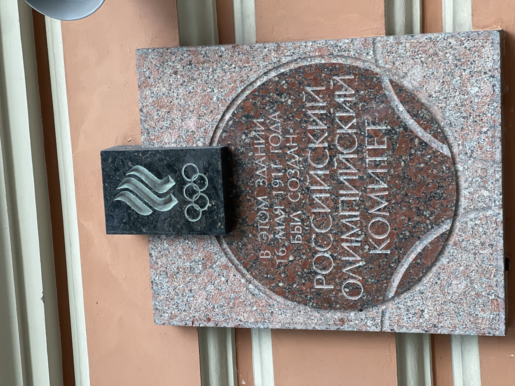 The image shows a commemorative plaque on the exterior wall of a building. The plaque is made of red granite with white and black speckles. At the top of the plaque, a black rectangular stone has the Olympic rings carved into it. Below the rings, text is carved into the granite. The text reads: "В ЭТОМ ЗДАНИИ 16 МАЯ 1911 ГОДА БЫЛ СОЗДАН РОССИЙСКИЙ ОЛИМПИЙСКИЙ КОМИТЕТ". This translates to: "In this building, on May 16, 1911, the Russian Olympic Committee was founded." The building has an orange, textured wall. The plaque is mounted on the wall, slightly below a white horizontal trim. The image is taken from a slightly low angle, looking up at the plaque.