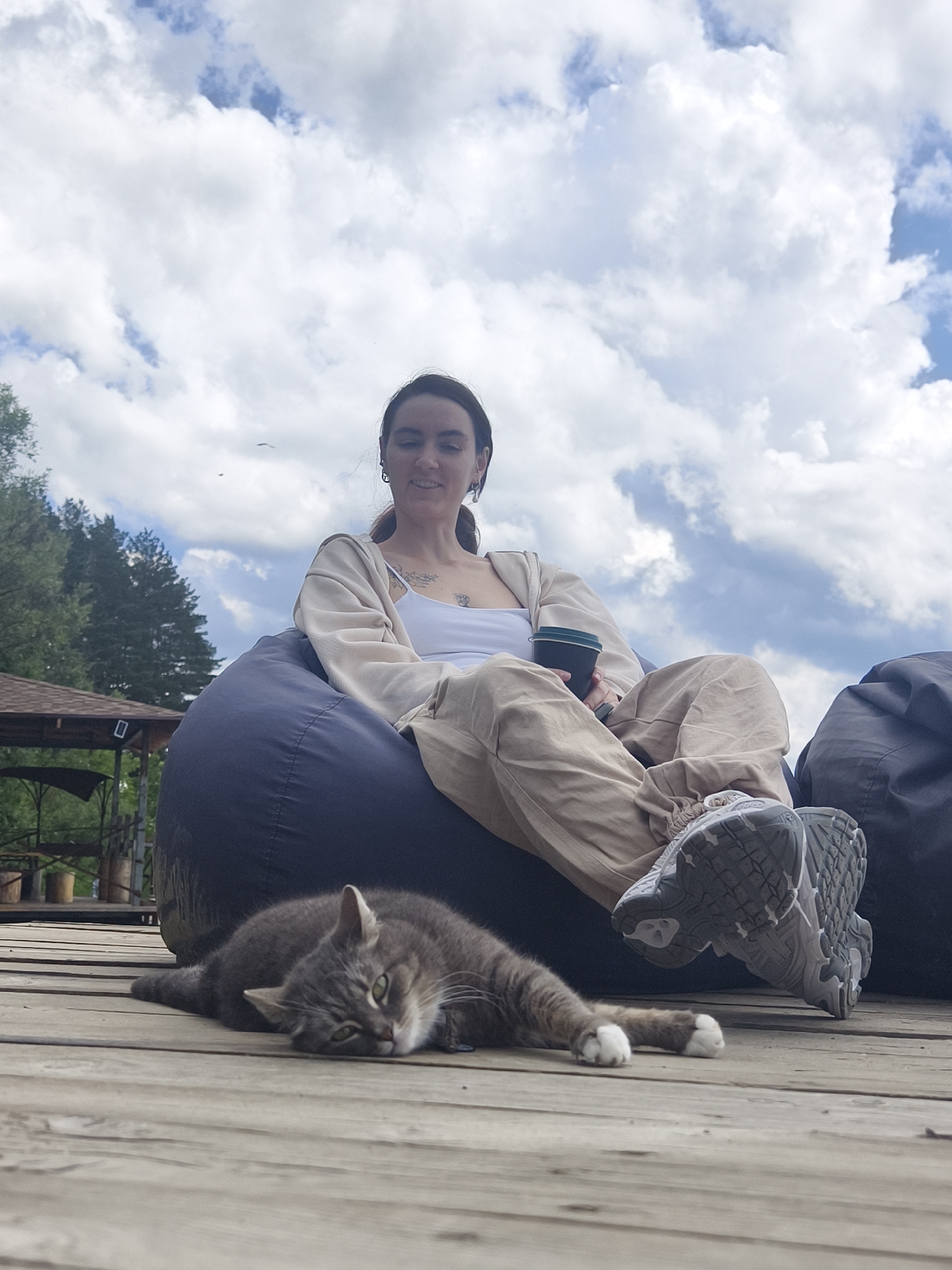 A young woman with dark hair tied back, wearing a white tank top, a beige open cardigan, and light-colored cargo pants, sits on a large, dark blue beanbag chair. She holds a dark cup in her right hand. Her feet are clad in grey athletic shoes. A grey tabby cat lies on its side on a wooden plank deck in front of the beanbag chair, its front paws extended towards the viewer. Behind the woman and to the left, a wooden structure with a peaked roof is visible, partially obscured by trees. The scene is outdoors under a sky filled with white, fluffy clouds. The location is the city of Онохино, Russia.