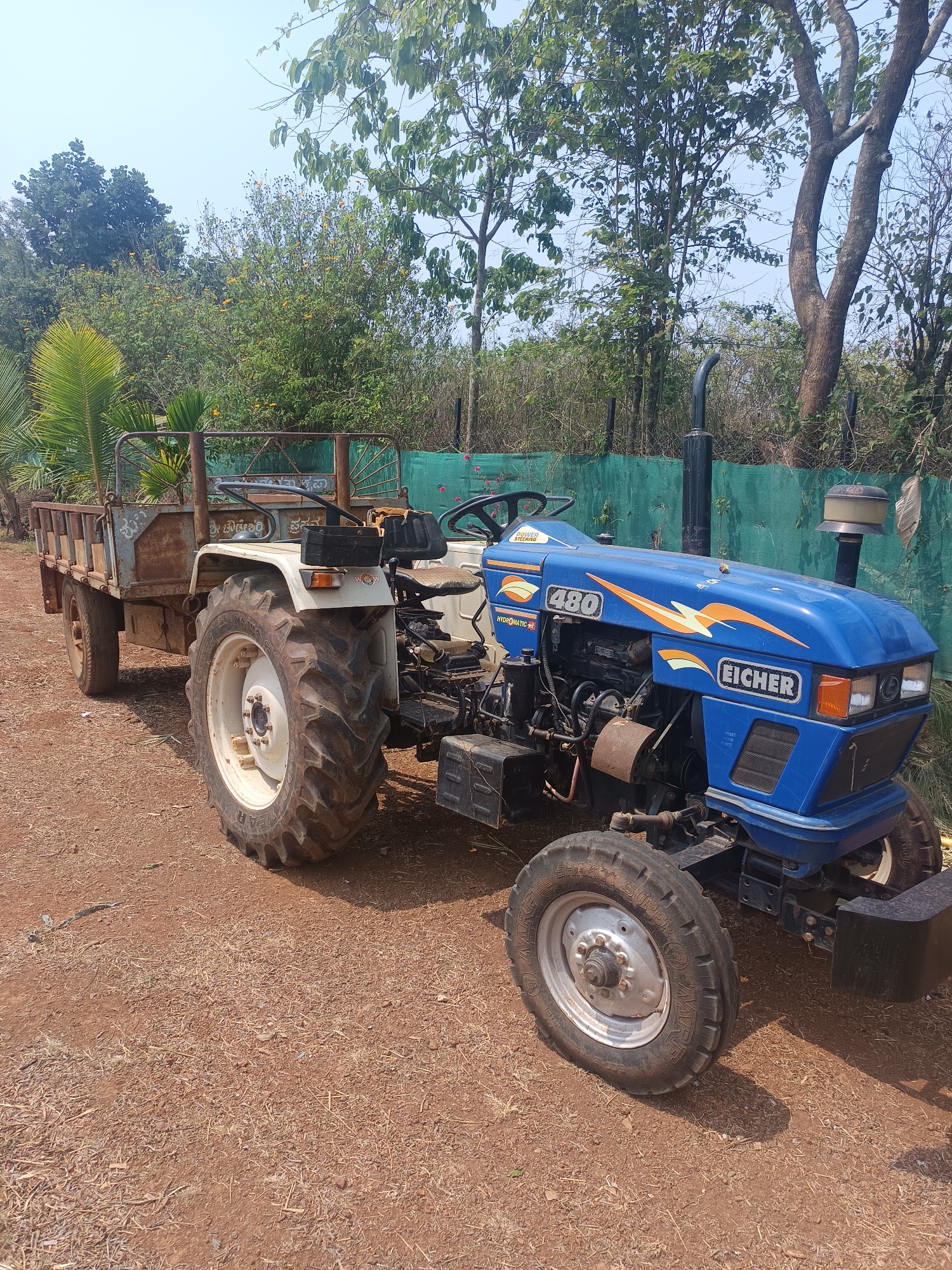 The image features a blue Eicher 480 tractor with an attached, rusty metal trailer, parked on a reddish-brown dirt path. The tractor, an older model, prominently displays "EICHER" on its hood, along with the model number "480" and decorative orange and white graphics. Additional text visible on the tractor includes "POWER STEERING" and "HYDROMATIC AF". The trailer is an open-top, two-wheeled unit showing significant rust and appears to have some text in a local language on its side, which is not clearly legible.
The setting is outdoors, under a clear, bright sky, indicating daytime. The ground is dry, reddish-brown earth mixed with small stones and organic debris. In the background, there is dense foliage consisting of various trees, including tall, leafy trees and some palm fronds on the left. A green mesh fence or netting runs across the background, separating the vehicle from the thicker vegetation. No people are present, and the tractor and trailer are stationary, suggesting they are either awaiting use or parked after work. The overall scene depicts a rural or agricultural environment, consistent with the location context of Dhundshi, India.