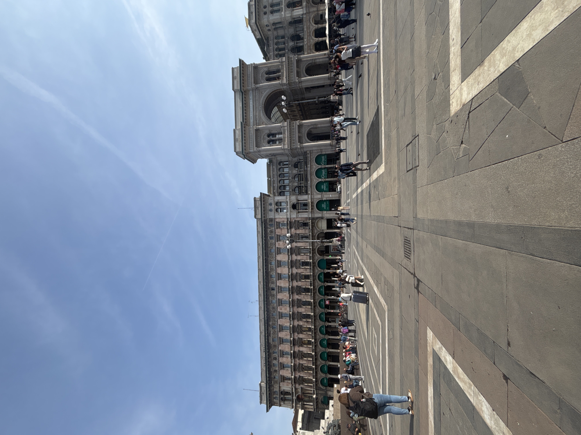 The image shows the Piazza del Duomo in Milan, Italy. In the background, the Galleria Vittorio Emanuele II is visible. The square is paved with grey stone and marked with a pattern of white squares and lines. A number of people are walking around the square. The sky is blue with some wispy clouds.
