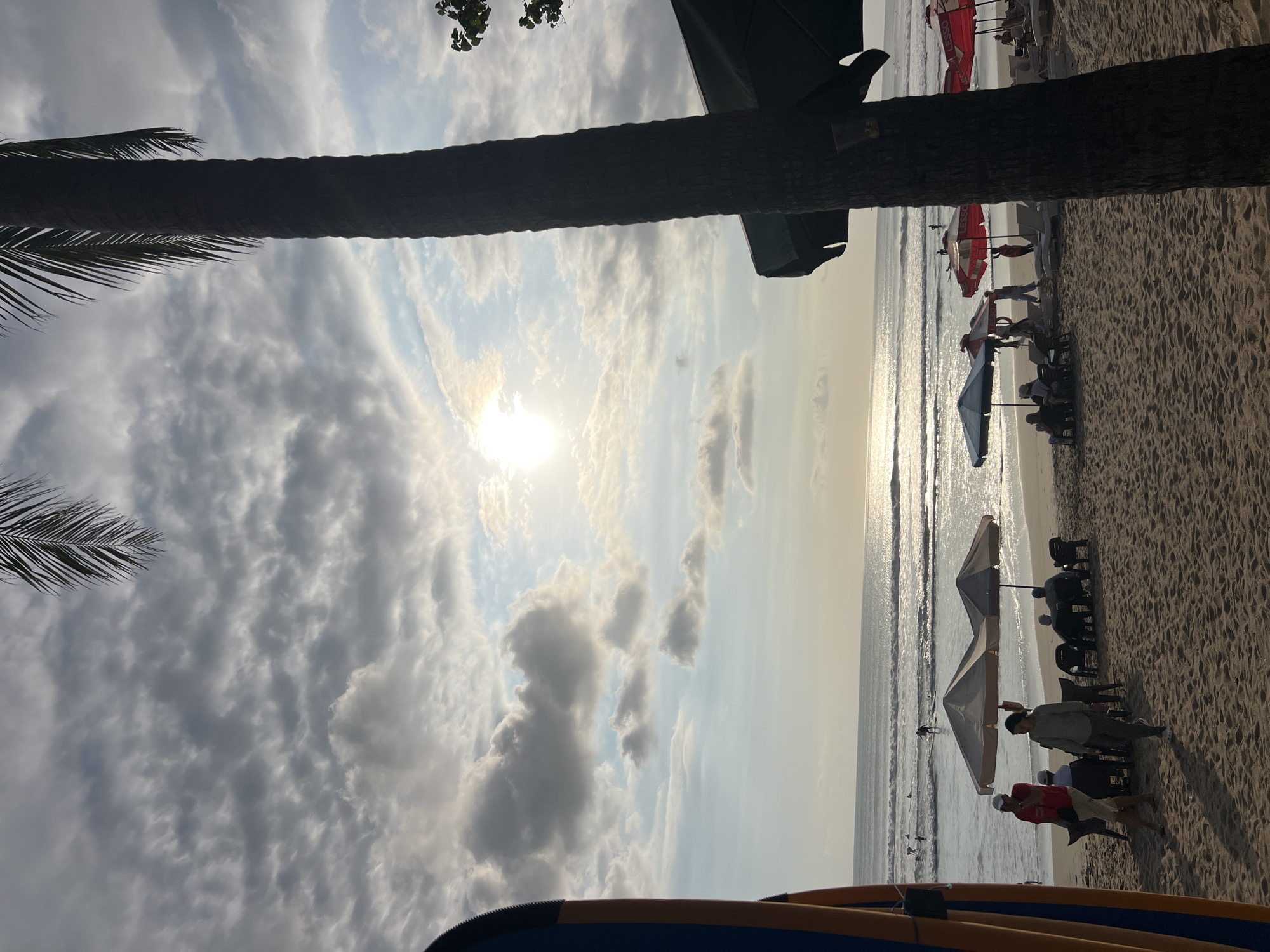 This is a medium-shot, eye-level, vertical photograph taken on the beach in Kuta, Indonesia, at sunset. The sun is low in the sky, casting a warm glow over the scene. The sky is filled with dramatic, scattered clouds that are illuminated by the setting sun. A tall palm tree dominates the right side of the frame, its dark silhouette contrasting with the bright sky. In the foreground, the sandy beach is textured with shadows.
Several beach umbrellas and lounge chairs are set up on the sand. Some people are seated under the umbrellas, appearing to relax. Others are walking on the beach or near the water's edge. In the distance, gentle waves roll onto the shore, and a few surfers are visible in the ocean. A surfboard is positioned on the far left edge of the image, leaning upright. The overall atmosphere is peaceful and serene, suggesting a late afternoon or early evening on a popular tourist beach.