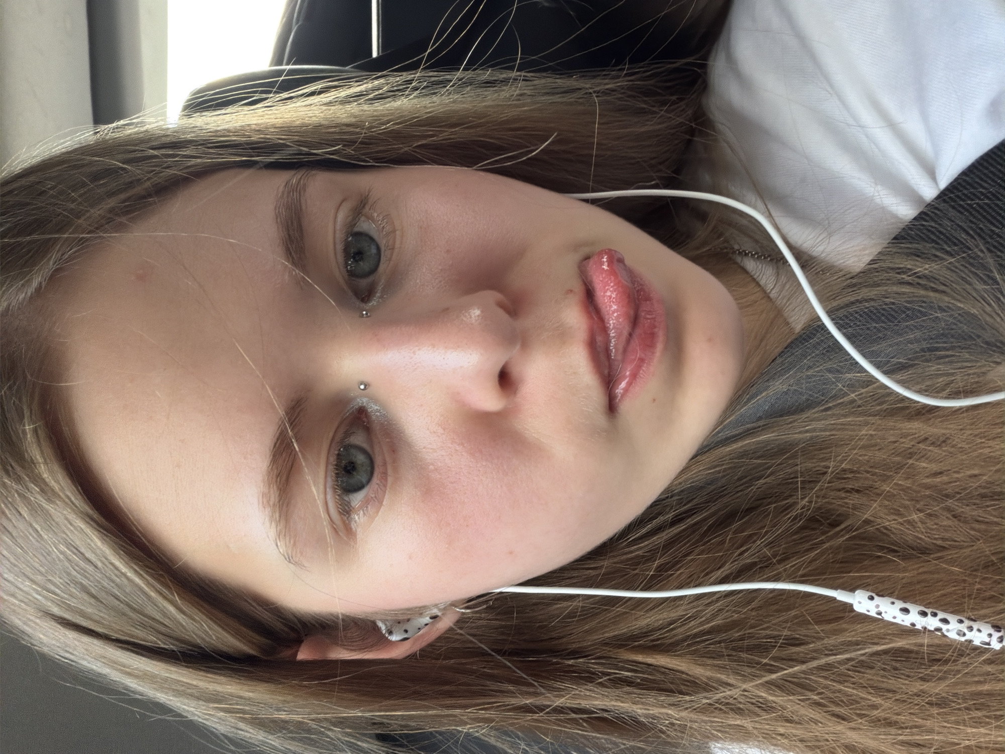 The image is a close-up, waist-up, selfie-style photograph of a young woman with long, light brown hair. She is wearing white earbuds with a black and white speckled pattern on the right earbud. She has a nose ring and an eyebrow piercing. Her tongue is sticking out, touching her lower lip. She is wearing a white t-shirt and a dark-colored seatbelt is visible across her chest. The background appears to be the interior of a vehicle, with a dark seat behind her and bright light coming from the right, suggesting it is daytime and possibly sunny. There is no discernible location in Moscow, Russia.