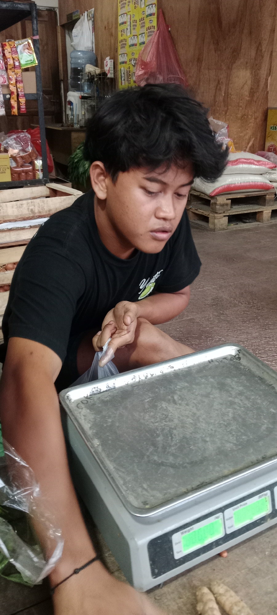 In Maguwoharjo, Indonesia, a young male wearing a black t-shirt is squatting in front of a metal weighing scale. He is holding a clear plastic bag, appearing to be preparing to place items on the scale. The background shows shelves stocked with various goods, including bottled water, packaged snacks, and large sacks of what appears to be rice. Wooden crates are visible, some containing produce. The overall environment suggests a market or a small shop setting. The lighting is moderate, illuminating the scene without harsh shadows.