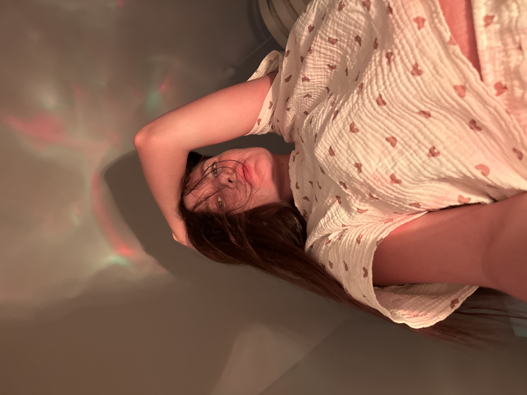 A young woman is seen in a selfie-style photo, tilted to the side. She has long, brown hair and green eyes. She wears a white, heart-patterned dress. The background appears to be a wall, with red and green light patterns. No information suggests this photo was taken in Belgrade, Serbia.