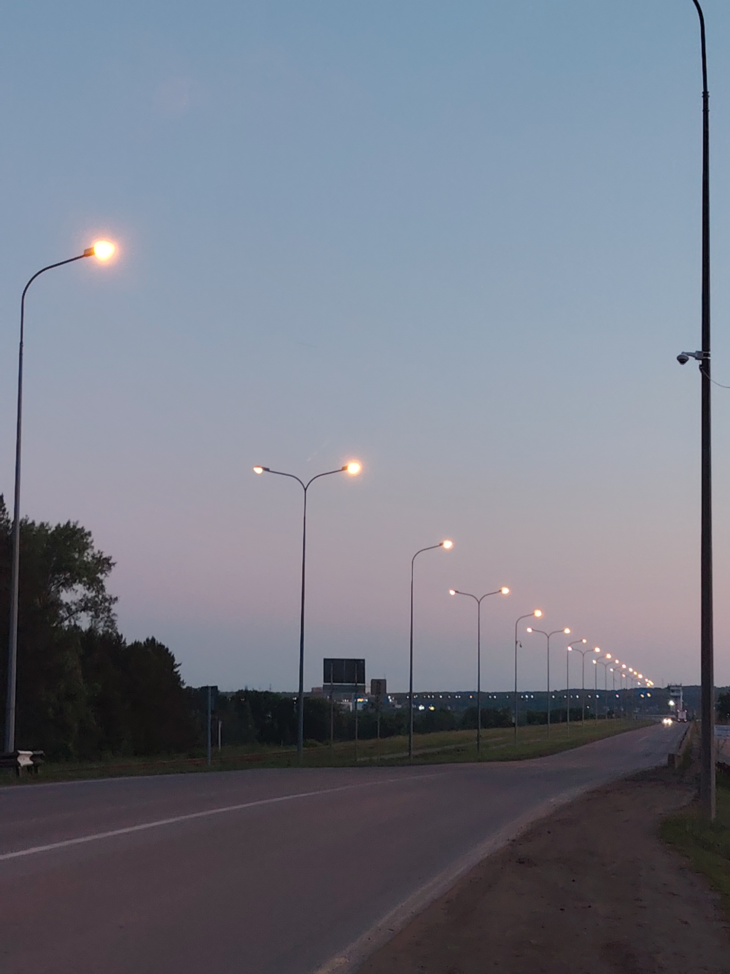 The image captures a serene, twilight scene along a multi-lane road in Chaikovsky, Russia. The sky, transitioning from a light blue at the top to a soft purple-pink near the horizon, indicates either dusk or dawn. Numerous streetlights line both sides of the road, casting a warm, orange glow and creating a sense of depth as they recede into the distance. The streetlights on the left are double-headed, while those on the right appear single. The leftmost streetlight is prominent in the foreground, brightly illuminated.
To the right, a tall streetlight pole features a security camera mounted about halfway up. The road surface is asphalt, with a clear white dividing line visible on the left lane. A dirt shoulder runs alongside the road on the right. Lush green trees and bushes border the road on the left, appearing dark against the fading light. In the mid-ground and background, more trees and hints of distant buildings or structures are visible. Far in the distance, what appears to be the headlights of a vehicle can be seen. No people or active events are visible, contributing to a quiet, peaceful atmosphere. No discernible text is visible in the image.