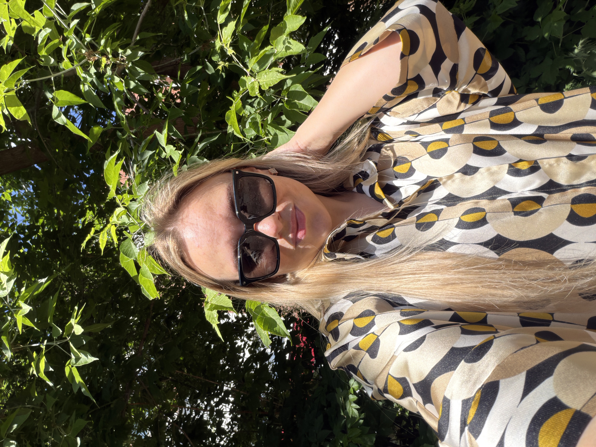The image depicts a female individual in an outdoor setting within the city of Votkinsk, Russia. She has long blonde hair and is wearing black rectangular sunglasses, with her mouth showing a subtle upward curve. She is dressed in a collared shirt featuring a repeating abstract geometric pattern composed of cream, mustard yellow, black, and white/light grey circular and semi-circular forms. Her left hand is raised, with fingers gently touching the hair near her left ear. The background is characterized by dense, vibrant green foliage from trees and bushes, suggesting a sunny day with bright highlights and shaded areas. Small sections of clear blue sky are visible through gaps in the leaves. A faint reddish-brown element, possibly a distant structure, is partially discernible through the foliage on the left.