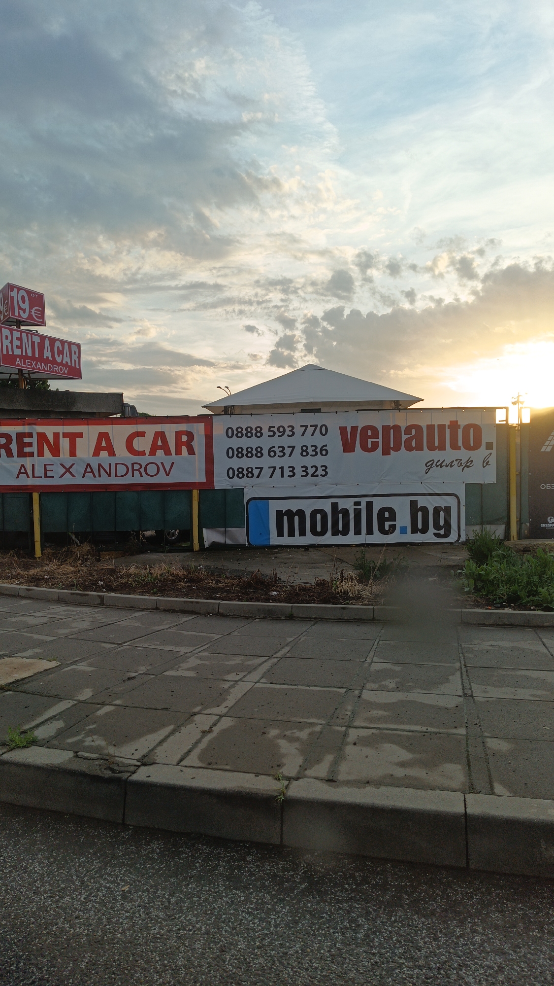The image shows a street scene in Burgas, Bulgaria, featuring advertisements for car-related businesses. On a green fence, banners advertise "RENT A CAR ALEXANDROV" alongside pricing. Contact phone numbers are displayed, along with the business name "vepauto дилър в" and "mobile.bg". The sky above is cloudy, with a hint of sunset. The foreground consists of a wet, tiled sidewalk and curb.