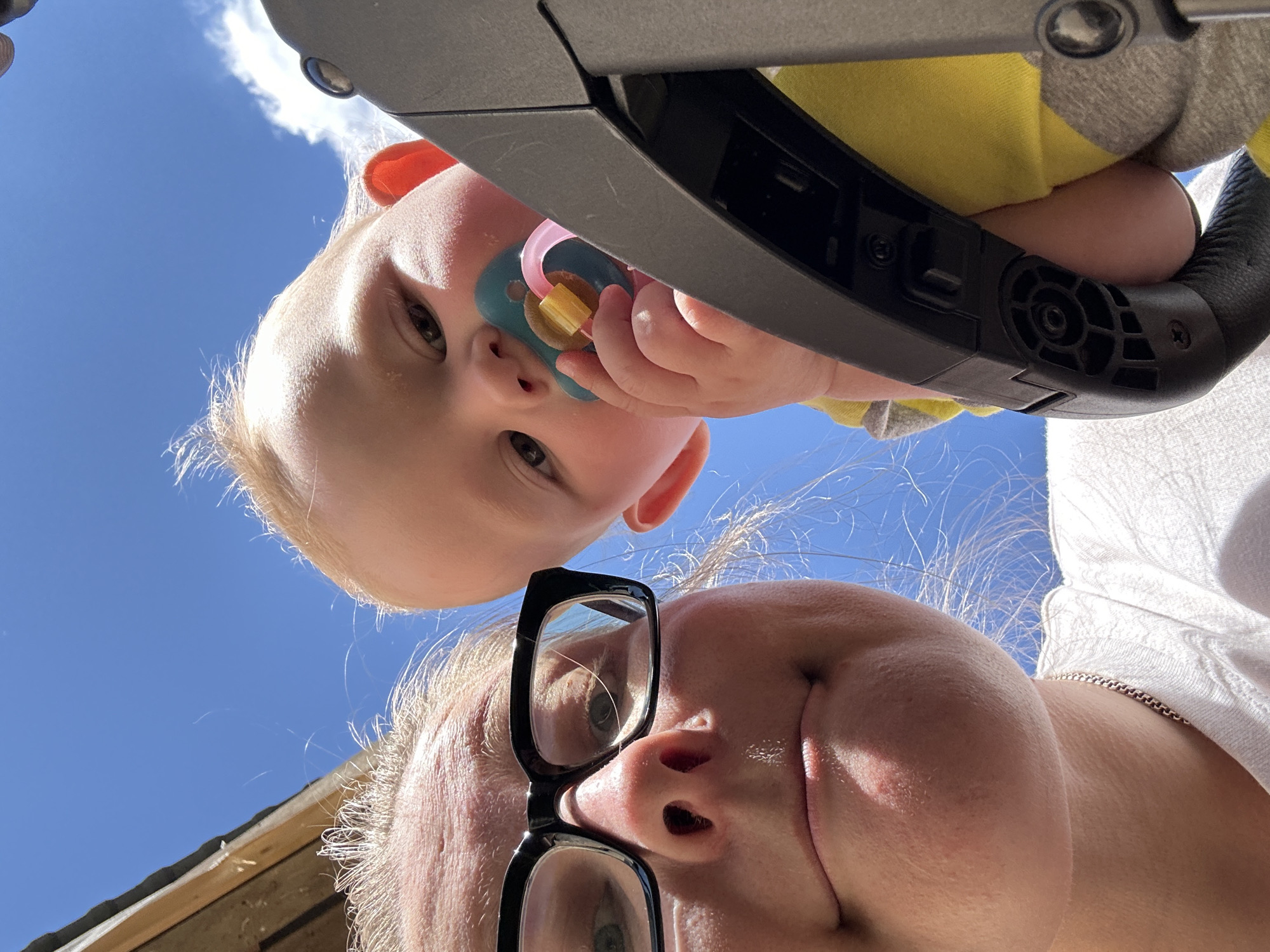 In Kalitsino, Russia, a low-angle, point-of-view shot captures a woman and a baby outdoors under a bright blue sky with scattered clouds. The woman, wearing black-rimmed glasses and a white t-shirt, is looking directly at the camera with a slight frown. Her face is partially obscured by the top of what appears to be a stroller. A baby, held by the woman, is looking towards the camera, with a pacifier in its mouth. The baby is wearing a yellow shirt with gray trim. The background shows a portion of a wooden structure on the left and the sky above. The lighting suggests it is daytime.