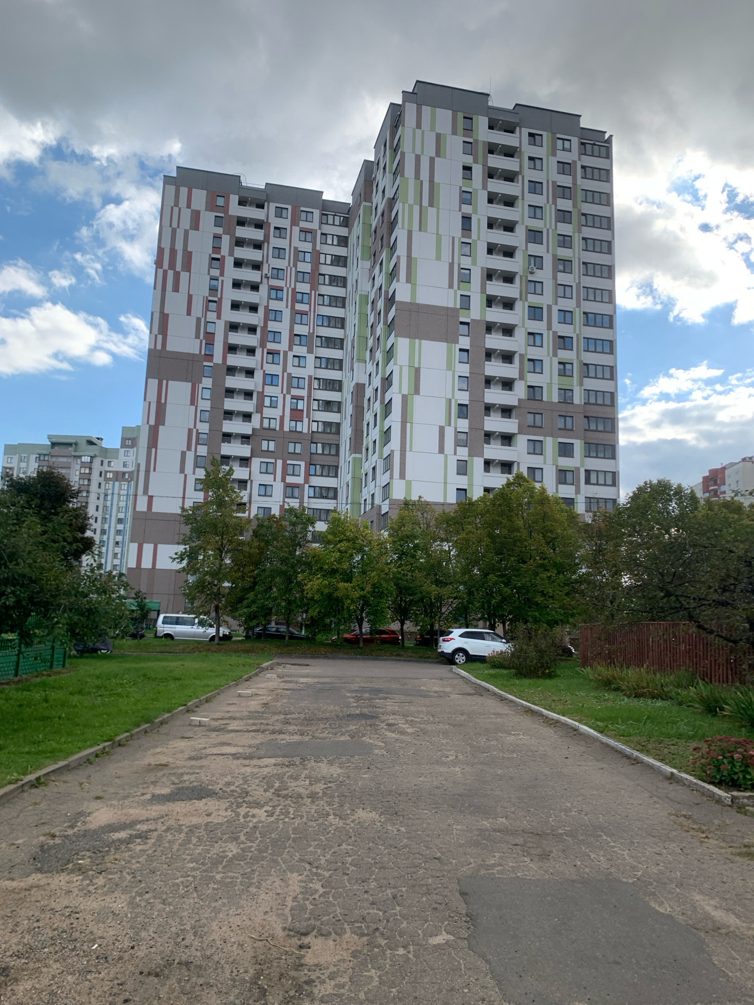 The image captures a view of two adjacent, modern high-rise apartment buildings in Minsk, Belarus. The buildings feature a striking facade with a patterned design incorporating shades of white, brown, and green. They are surrounded by trees and greenery, with a paved area in the foreground, possibly a road or parking lot. The sky is partly cloudy with patches of blue sky visible. Several cars are parked in the vicinity of the buildings. The scene appears to be a residential area within the city.