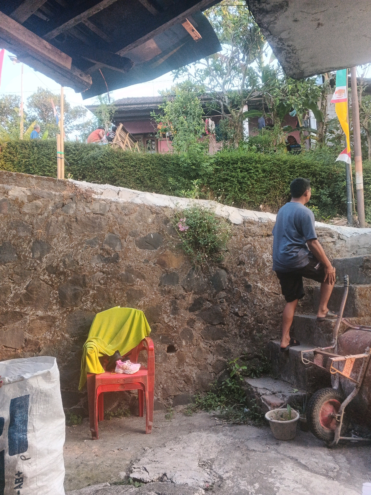The image depicts an outdoor scene in Keji, Indonesia. In the foreground, a white, woven bag with blue markings stands on a rough concrete surface. To its right, a red plastic chair is draped with a bright yellow cloth, and a pair of pink children's shoes rests on the seat. Behind the chair, a weathered stone wall forms the backdrop.
A young man, wearing a dark grey t-shirt and black shorts, is ascending a set of concrete stairs to the right of the frame. His back is to the camera, and he is looking towards the top of the stairs. At the base of the stairs, a small, weathered clay pot contains a green plant. To the far right, a rusty wheelbarrow is partially visible.
In the mid-ground, a dense green hedge lines the top of the stone wall. Beyond the hedge, a building with a red roof and a dark-colored facade is visible, surrounded by lush vegetation. Several figures are present in the background, some near the building and others further up a slope, partially obscured by trees. A flagpole with a red, white, and green flag is also visible. The overall impression is of a rural or semi-rural setting, possibly a residential area or construction site, with signs of ongoing activity.
