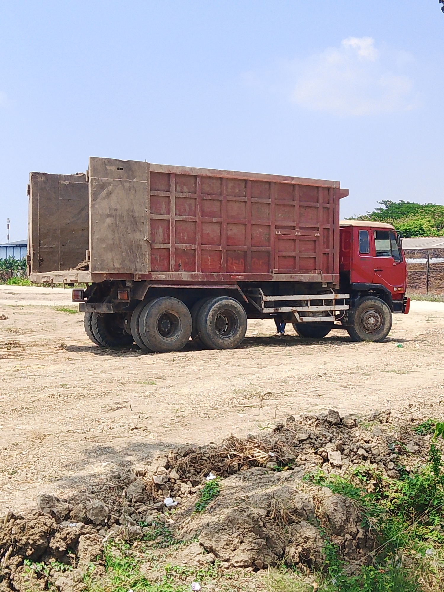 In Bermi, Indonesia, a large, weathered red dump truck sits parked on a dusty, unpaved lot under a bright blue sky with scattered clouds. The truck has a dual rear axle and a large, open bed that appears to be made of metal and wood, with some debris visible inside. The truck is parked at an angle, with its front facing towards the right side of the frame. In the background, there are some trees, a fence, and a building structure. The ground is covered in dry dirt and some piles of soil and vegetation are in the foreground. The overall scene suggests an industrial or construction site.