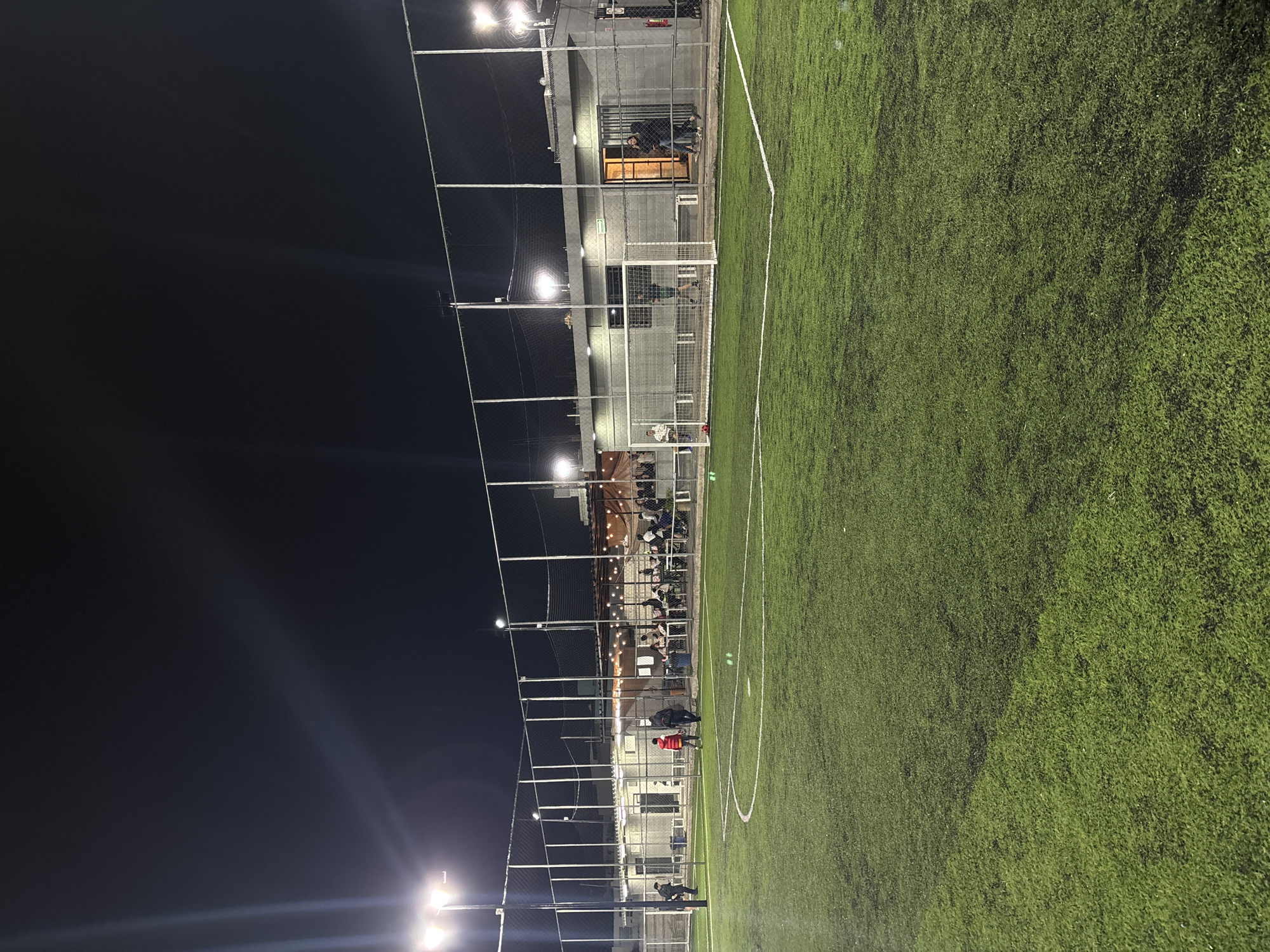 The image shows a soccer field at night in San Bernardino Tlaxcalancingo, Mexico. The field is illuminated by stadium lights, and the artificial turf is visible. A fence surrounds the field, and a building runs parallel to it. Spectators are visible behind the fence and along the building. There are a few people visible on the field.
