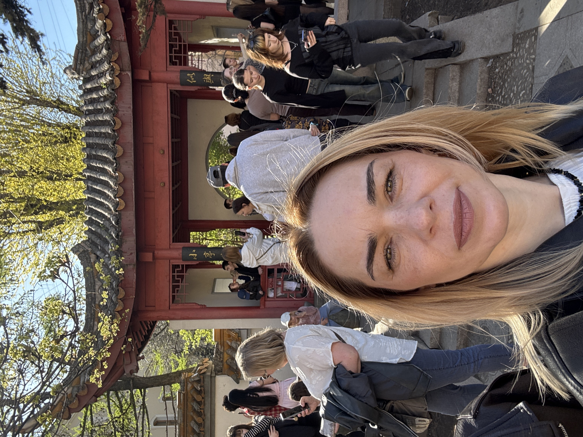 The image is a selfie of a fair-skinned woman with blonde hair. She is wearing a dark jacket with a white top underneath. Behind her, a crowd of people is gathered near a traditional Chinese-style pavilion. The pavilion has red columns and a grey tile roof. There are signs with Chinese characters on the pavilion. In the background, there are trees with budding green leaves, suggesting it is springtime. The location is Friendship Garden in Saint Petersburg, Russia.