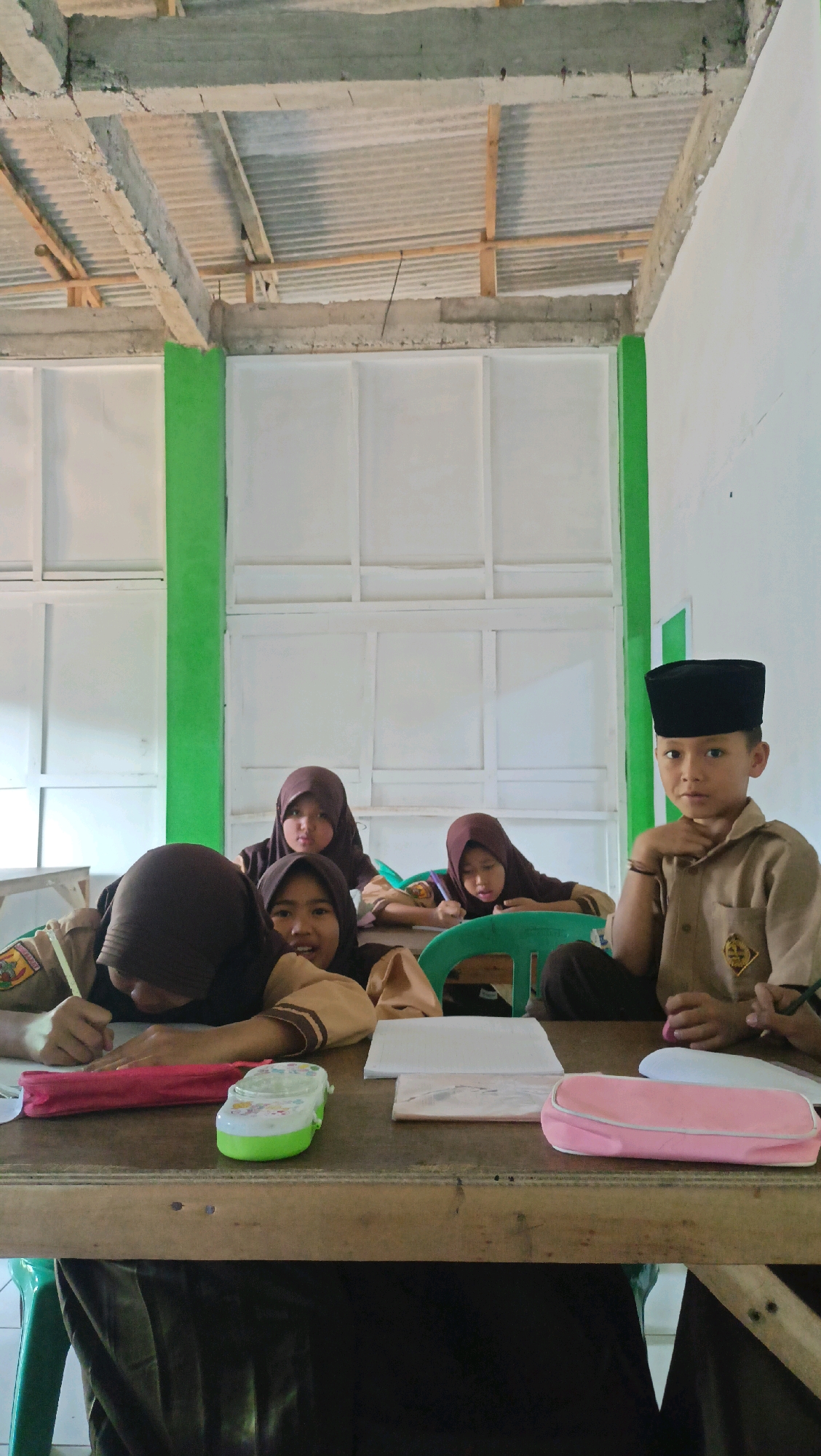 This is a medium-shot, portrait-oriented photograph of a group of five Indonesian schoolchildren seated at a wooden table, presumably in a classroom in Tugumukti, Indonesia. The room appears unfinished, with exposed concrete beams and corrugated metal roofing visible in the background, partially obscured by a green-painted beam. The walls are white and feature a grid-like pattern.
In the foreground, on the table, are several school supplies. A red pencil case, a green and white pencil box, and a pink pencil case are present. Two sheets of paper are also visible, one appearing to be a workbook.
Five children, all wearing brown uniforms and headscarves (hijabs), are the subjects.
On the left, a child in the front row is leaning over the table, writing with a pencil. To their right and slightly behind, another child is also looking down at the table, possibly writing.
Further back, two more children are seated, one in the center and one to the right. They are also looking down, seemingly engaged in schoolwork.
On the far right, a boy wearing a black peci (cap) and the same brown uniform is looking directly at the camera, resting his chin on his hand. He appears to be the oldest child in the group.
The lighting is natural and somewhat diffused, suggesting it is daytime. The overall impression is a candid snapshot of children engaged in learning in a basic educational setting.