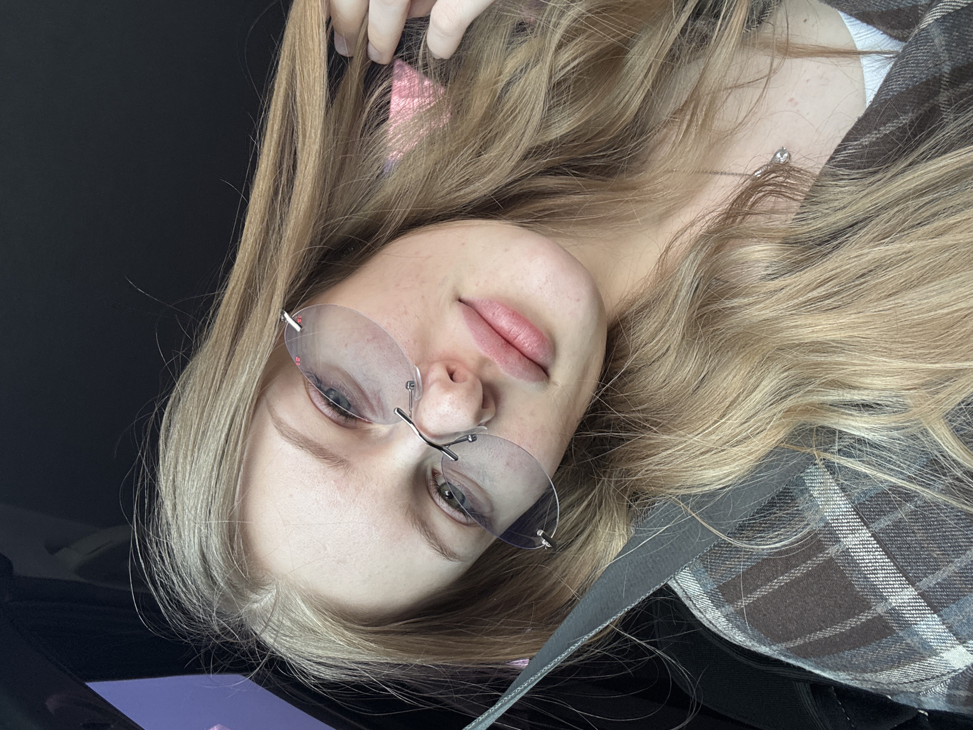 A young Caucasian woman with long blonde hair and fair skin is pictured indoors in a portrait-style photograph. She is wearing rimless eyeglasses and a plaid shirt or jacket. A seatbelt is visible across her body, suggesting she might be in a car. The background is dark, possibly the interior of a vehicle. There is no indication of the city of Perm, Russia in the provided image.
