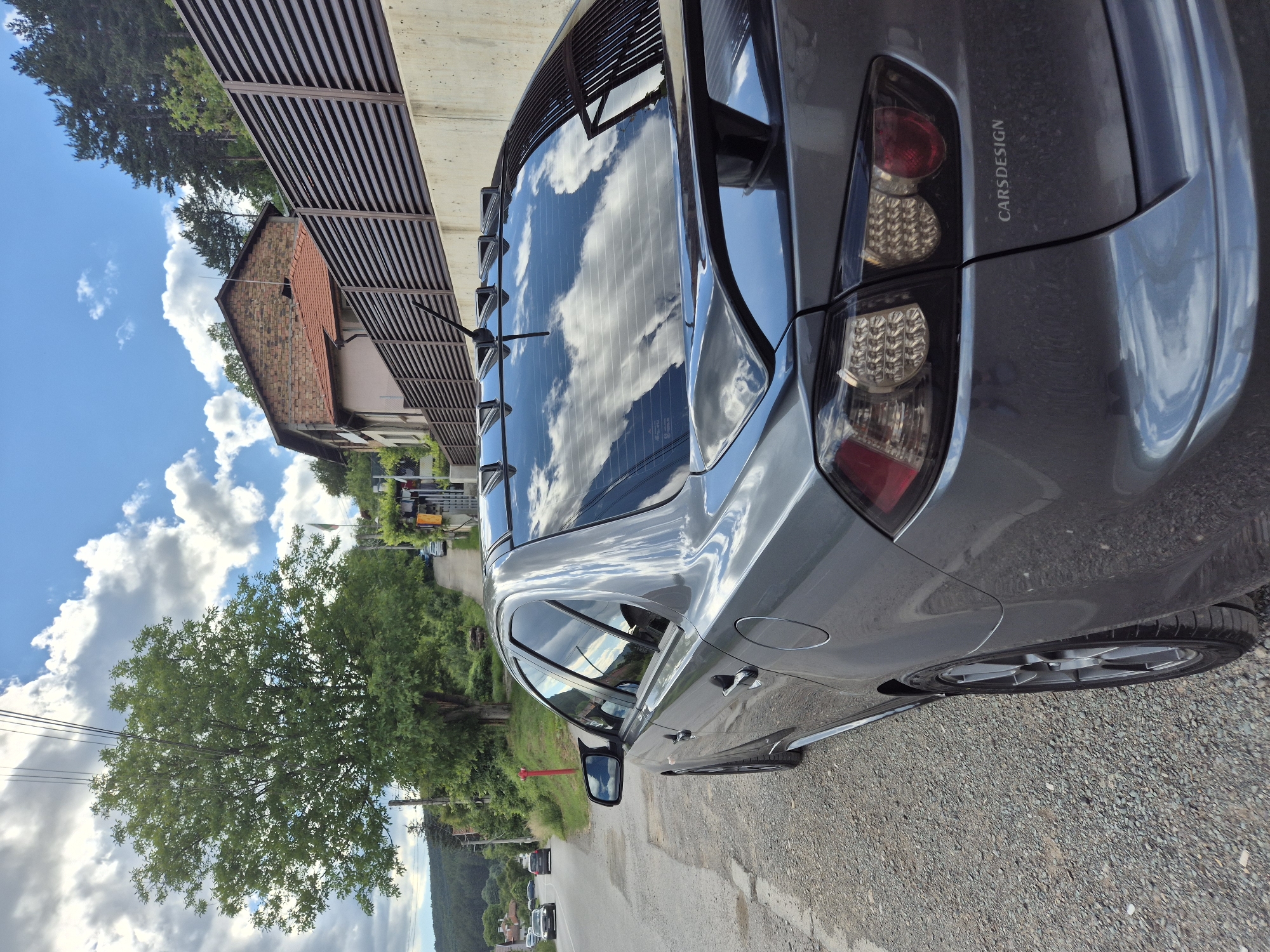 A grey sedan is parked on the side of a rural road in Iskrets, Bulgaria, under a partly cloudy sky. The car, positioned at an angle facing away from the viewer and towards the right, has its rear visible, showcasing tinted windows with reflections of the sky and surrounding trees. A rear spoiler is mounted on the trunk. The road is gravel, and to the right of the car, there is a concrete wall topped with a dark, slatted fence. Behind the fence, a two-story building with a red-tiled roof and brick facade is visible, situated on a hillside covered with trees. In the background, the road winds uphill, with other vehicles and houses visible further down the street. A large tree with green foliage stands prominently on the left side of the road, and power lines extend across the sky.