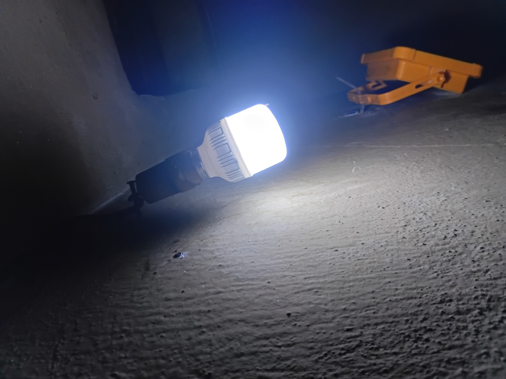 The image shows a white LED light bulb screwed into a black fixture, hanging from the ceiling in a dimly lit area. The bulb is illuminated, casting a bright white light. To the right of the bulb, an orange clamp-like object is attached to the wall with what appear to be zip ties. The wall itself has a rough, textured surface, with visible pores and streaks. The environment appears to be an indoor space, possibly a workshop or storage area, given the utilitarian nature of the lighting and the presence of the clamp. The scene is likely located in Nkpor, Nigeria, as specified.