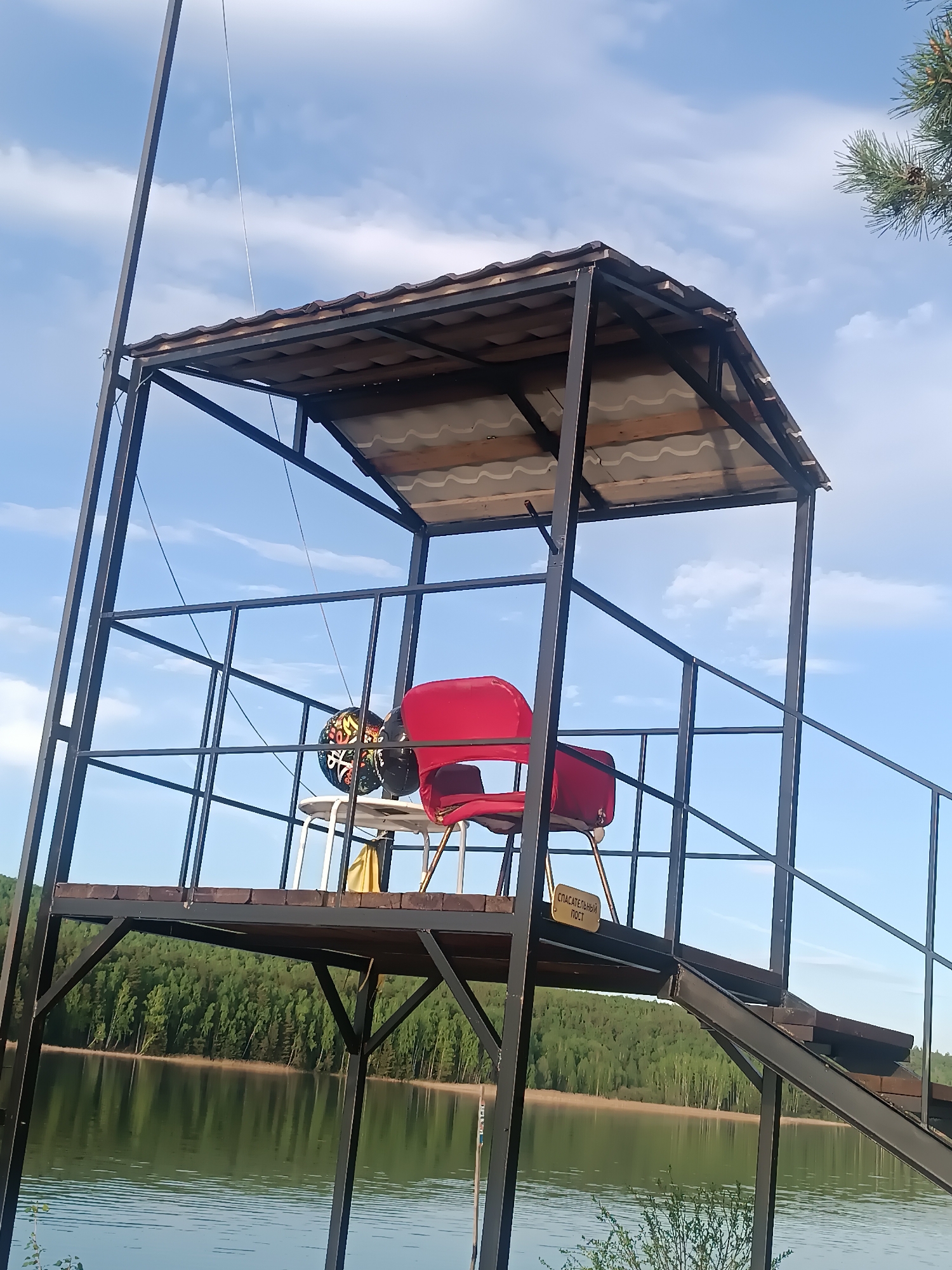 The image shows a metal lifeguard tower by a body of water, possibly a lake, surrounded by trees. The tower has a roof, a deck with railings, and a staircase leading up to it. On the deck, there are two red chairs and a small white table with balloons and other objects on it. A yellow sign with writing in a foreign language is attached to the tower. The location is unknown, Unknown.