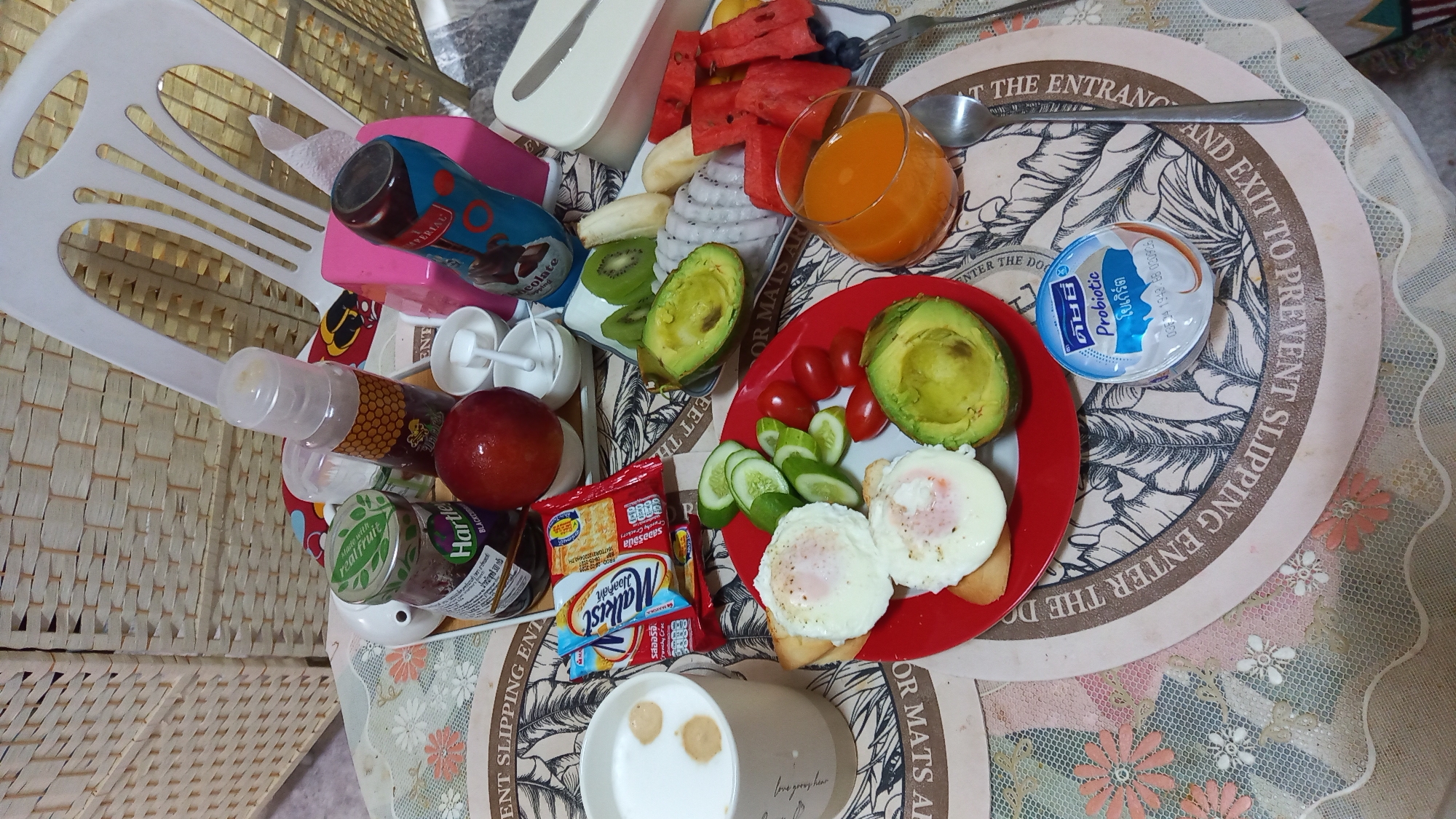 The image displays a breakfast spread on a table, likely in Ko Pha-ngan District, Thailand. The scene is dominated by a variety of food and drink items. In the foreground, a red plate holds two poached eggs on toast, accompanied by sliced cucumbers, cherry tomatoes, and half an avocado. To the left, a white mug contains a milky beverage with frothy accents. A glass of orange juice is positioned to the right of the plate.
Further back on the table, a platter features a selection of fruits, including sliced kiwi, bananas, dragon fruit, and watermelon. A jar of Hartley's jam and a bottle of what appears to be chocolate syrup are also visible. A pink tissue box and a white plastic container are present towards the top right of the frame. A small container of "Probiotic" yogurt is situated in the lower right quadrant. The table is covered with a patterned placemat. The background shows a woven basket-like material and a portion of a white plastic chair. The lighting suggests an indoor setting, possibly a home or a casual dining area. No people are visible in the image.