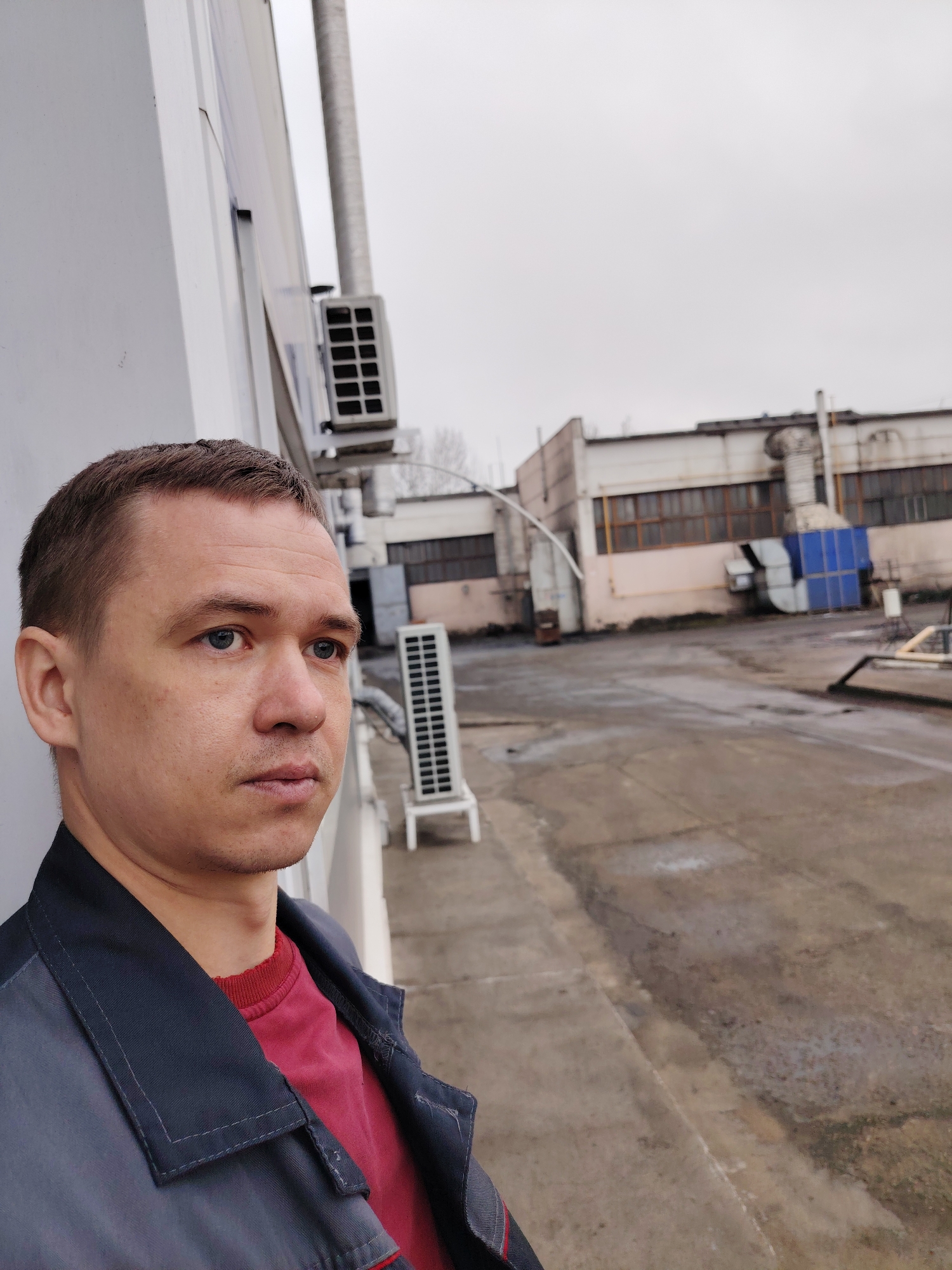 A medium shot depicts a man in the foreground, slightly left of center, wearing a red shirt beneath a gray or blue work jacket. He gazes to the right. The background features an industrial outdoor area. An air conditioning unit is visible above and to the right of the man’s head, and another similar unit stands on the ground further back. The ground appears to be concrete or asphalt. In the distance, there is a long, low building with multiple windows and pipes protruding from the roof. The sky is overcast. The location is Noviy, Russia.