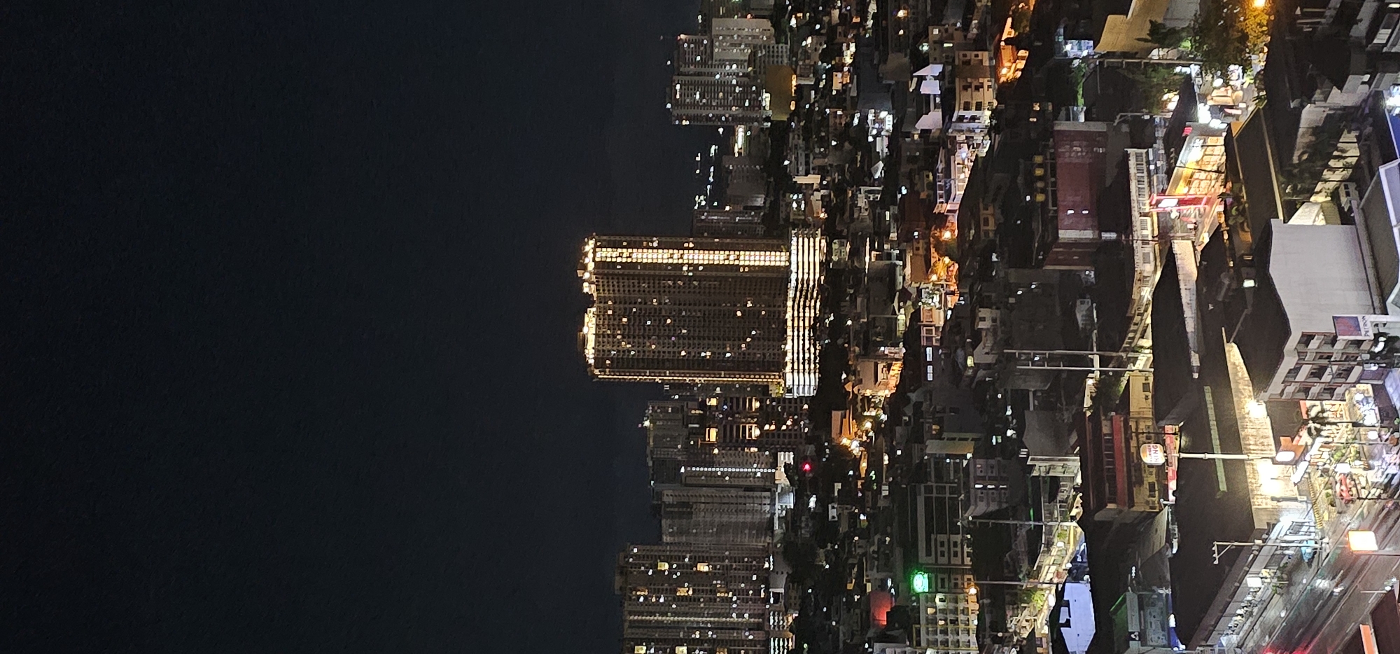 A high-angle, nighttime, long-shot captures a dense urban landscape in Makati, Philippines. The city is illuminated by a multitude of artificial lights from buildings and streets. In the foreground, the scene is dominated by various buildings, including a Burger King establishment with its distinctive signage, and a building with "PETRON" branding. The middle ground features a cluster of lower-rise structures, some with visible lights indicating occupancy. In the background, towering skyscrapers with illuminated windows pierce the dark sky. The overall impression is one of a bustling, brightly lit metropolis at night.