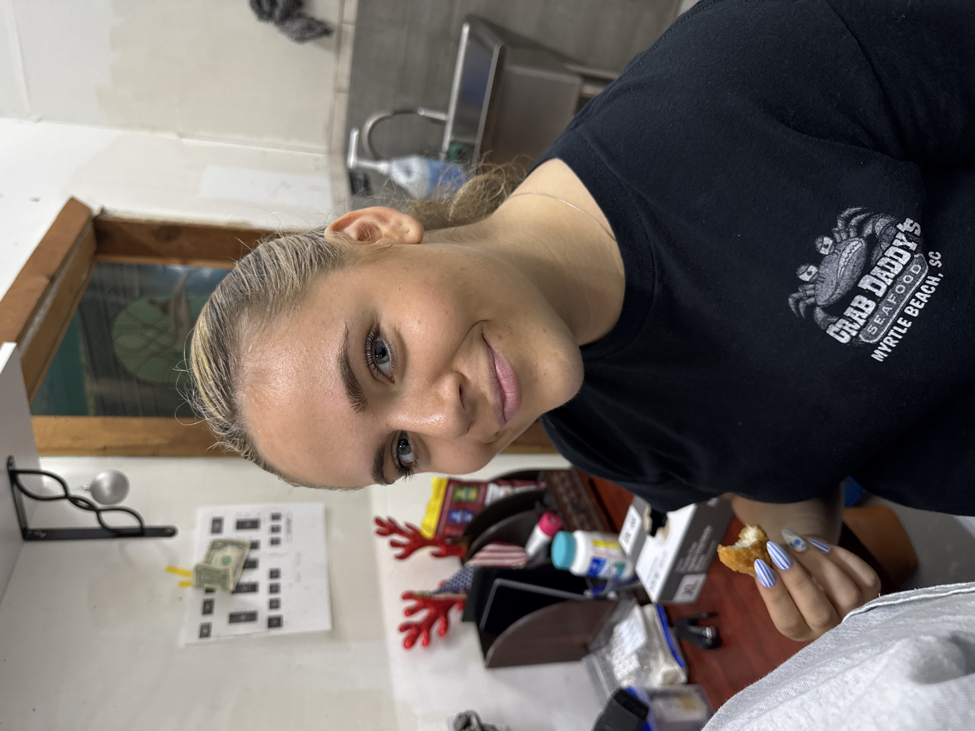A woman with blonde hair and blue eyes is wearing a black t-shirt with a "Crab Daddy's Seafood" logo on it, indicating the location is likely in Myrtle Beach, SC. She is holding a piece of food, possibly chicken, in her right hand, which has blue and white striped nails. She is smiling slightly at the camera. Behind her, there's a cluttered counter with various items, including a dollar bill, office supplies, and holiday decorations. The background suggests a kitchen or office area, possibly in a commercial setting.