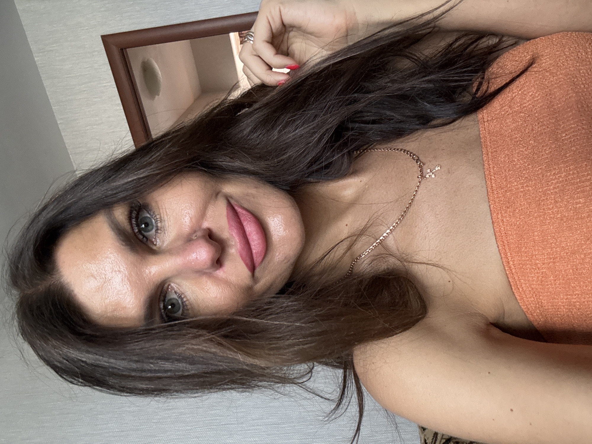 The image is a selfie of a woman with long, dark brown hair and blue eyes. She is wearing an orange strapless top and a gold necklace with a cross pendant. She has makeup on, including pink lipstick. Her skin appears to be tanned and has some texture. In the background, there is a wall with a textured pattern and a wooden-framed mirror. There is no information about the location being Dubovaya, Russia in the image.