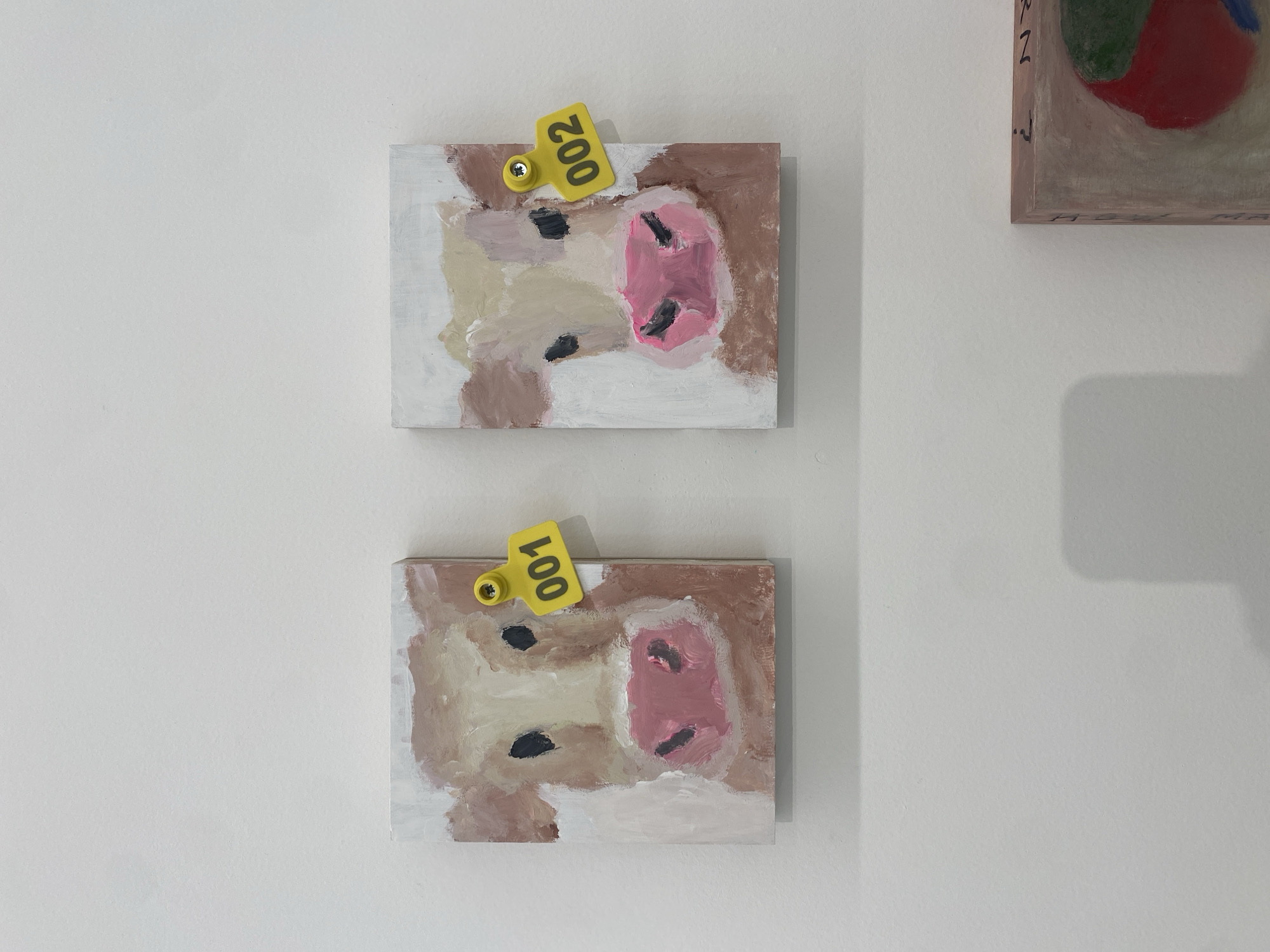 Two paintings of a cow's face, each adorned with a yellow tag, hang on a white wall. The top painting features a tag labeled "200," while the bottom one displays "100." The style of the paintings is abstract and rudimentary. The adjacent painting appears to have colorful balls and is painted in pastels. The location is unknown.