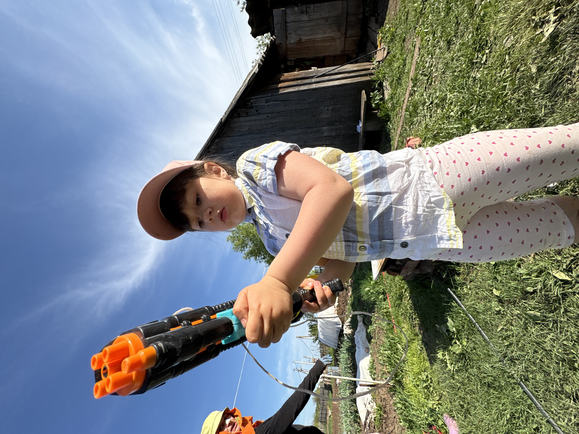 A young girl holds a black and orange toy gun pointed toward the camera. She wears a pink cap, a striped yellow and blue shirt, and white leggings with pink hearts. The scene is outdoors with grass, plants, and a shed in the background. A person in a black jacket and a yellow hat is partially visible below the girl. The sky is blue with light clouds. The scene is in the city of Nizhnyaya Chermoda, Russia.