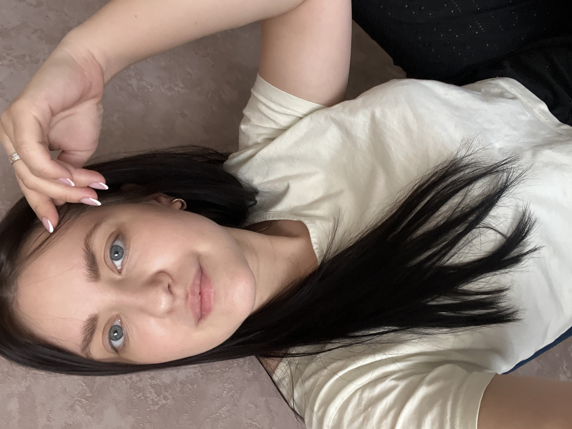 This image depicts a female subject indoors in the city of Марковский, Russia. The subject is a young adult woman with long, straight, dark brown hair and blue eyes. She has a fair complexion and a neutral facial expression, looking directly forward. She is wearing a light-colored, possibly off-white, short-sleeved t-shirt. Dark-colored fabric, likely pants, is visible on the lower right side of the frame. A gold hoop earring is present in her left ear. Her right hand is raised, with fingers positioned near her forehead and hairline, and a multi-banded ring is visible on the fourth finger of this hand. Her fingernails exhibit a French manicure style with white tips. The background consists of a textured, light brown or beige wall surface.