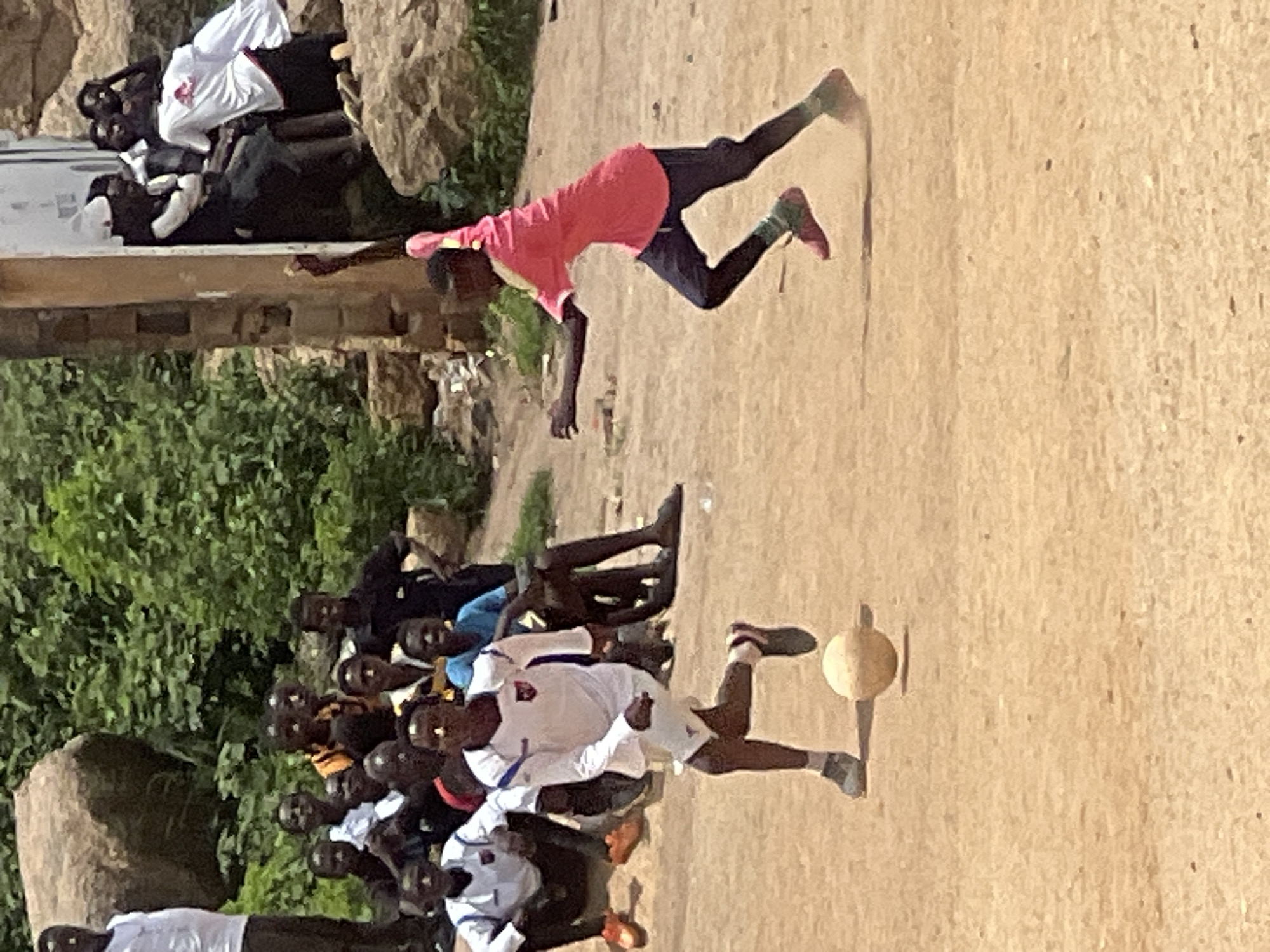 In the city of Jos, Nigeria, this photograph captures a lively outdoor football match. The scene unfolds on a dusty, uneven pitch, with onlookers gathered in the background. The backdrop consists of a mix of natural elements, including large rocks and lush green vegetation, alongside some rudimentary structures suggesting a semi-urban or rural setting.
The foreground is dominated by two young boys actively engaged in the game. One boy, dressed in a white long-sleeved jersey and white shorts, is in the process of kicking a light-colored football. His stance and the position of the ball indicate he is either passing or shooting. The other boy, wearing a bright pink t-shirt and dark trousers, is running dynamically, seemingly chasing the ball or positioned to receive a pass. His movement suggests speed and agility.
In the mid-ground and background, several other individuals, predominantly young males, are observing the match. They are positioned on the side of the pitch and appear to be spectators, some sitting, others standing. Their attire varies, with some in casual wear and others in what might be team jerseys. The overall atmosphere appears energetic and communal, typical of a local sporting event. The lighting suggests it is daytime, likely a sunny afternoon.