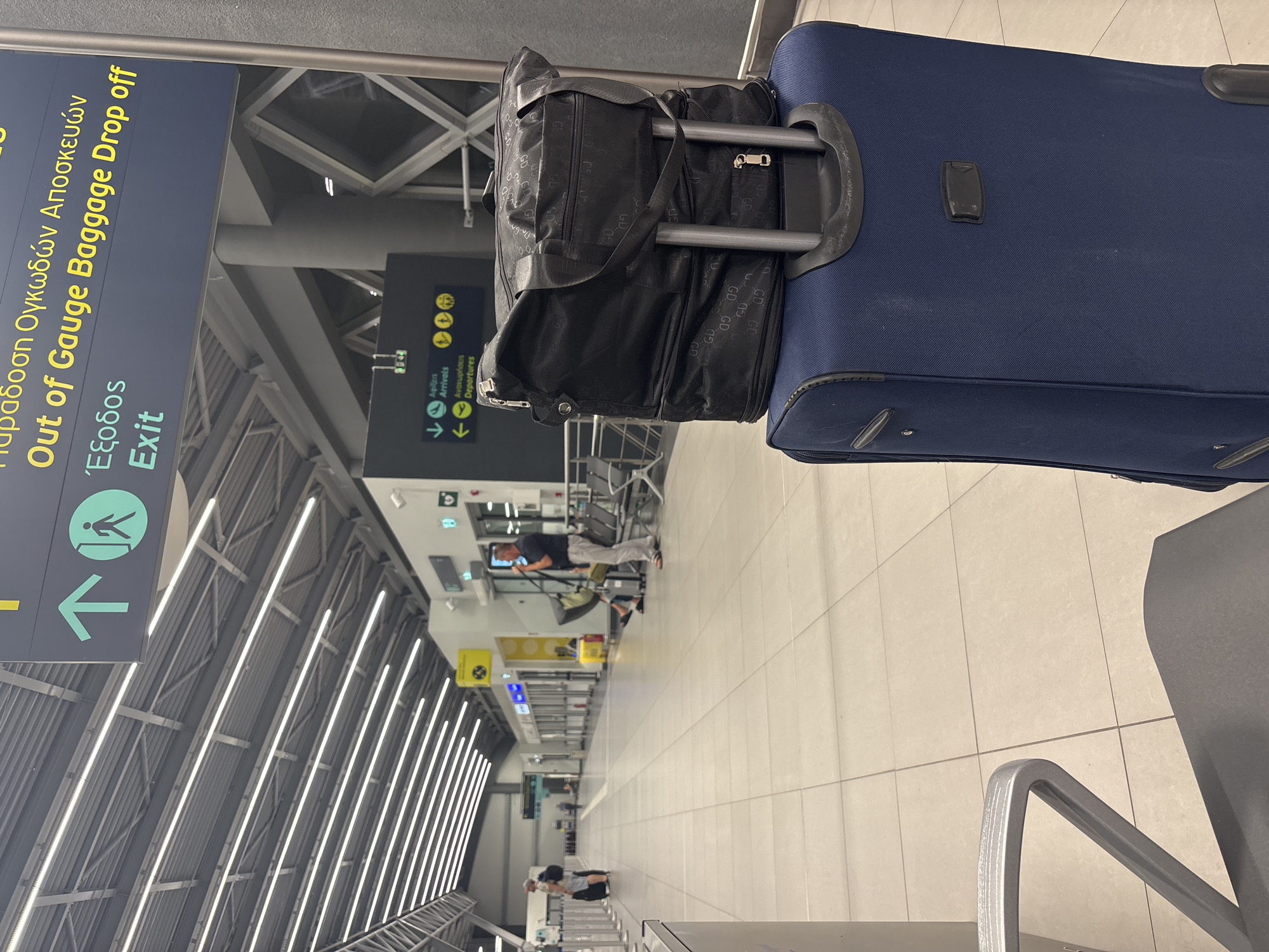 In Thermi, Greece, at the Thessaloniki Airport (SKG), a dark blue, two-wheeled suitcase stands in the foreground. A black duffel bag, featuring a repeating "GD" pattern and a textured finish, is placed on top of the suitcase. The duffel bag has a shoulder strap and multiple zippered compartments.

In the background, the airport's interior is visible. Signs overhead indicate "Out of Gauge Baggage Drop off" and "Exit" in both Greek and English, with accompanying directional arrows and icons. Further down the concourse, other travelers are seen with their luggage. One person is standing near a check-in counter, while another is seated, looking at their phone. The environment is brightly lit by overhead fluorescent lights and features a polished tiled floor and a metallic ceiling structure.