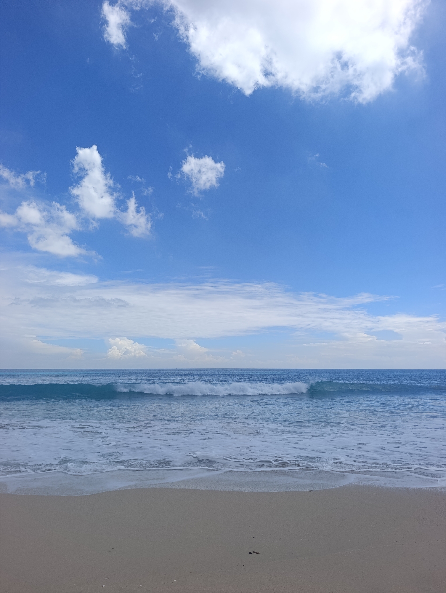 The image presents a tranquil seascape featuring a sandy beach, the ocean, and a blue sky, likely captured in Kuta Selatan, Indonesia. In the foreground, a stretch of light beige sand appears mostly untouched, with gentle foamy white waves receding across its surface.
The mid-ground is dominated by the clear blue-turquoise ocean. Several waves are visible, with a prominent one actively breaking and forming a white crest as it rolls towards the shore. In the deeper water further out, tiny dark specks suggest the presence of people, possibly swimmers or surfers, enjoying the water, though they are very small and indistinct.
The upper two-thirds of the frame are filled with a bright, clear blue sky. Scattered white cumulus clouds of various sizes adorn the sky, ranging from small, fluffy formations to larger, more expansive clusters. Some wispy cloud formations are also visible closer to the horizon.
The overall impression is of a beautiful, sunny day at the beach, characterized by a calm and natural atmosphere. The absence of prominent human activity in the immediate foreground enhances the sense of peaceful solitude. No discernible text is visible in the image.