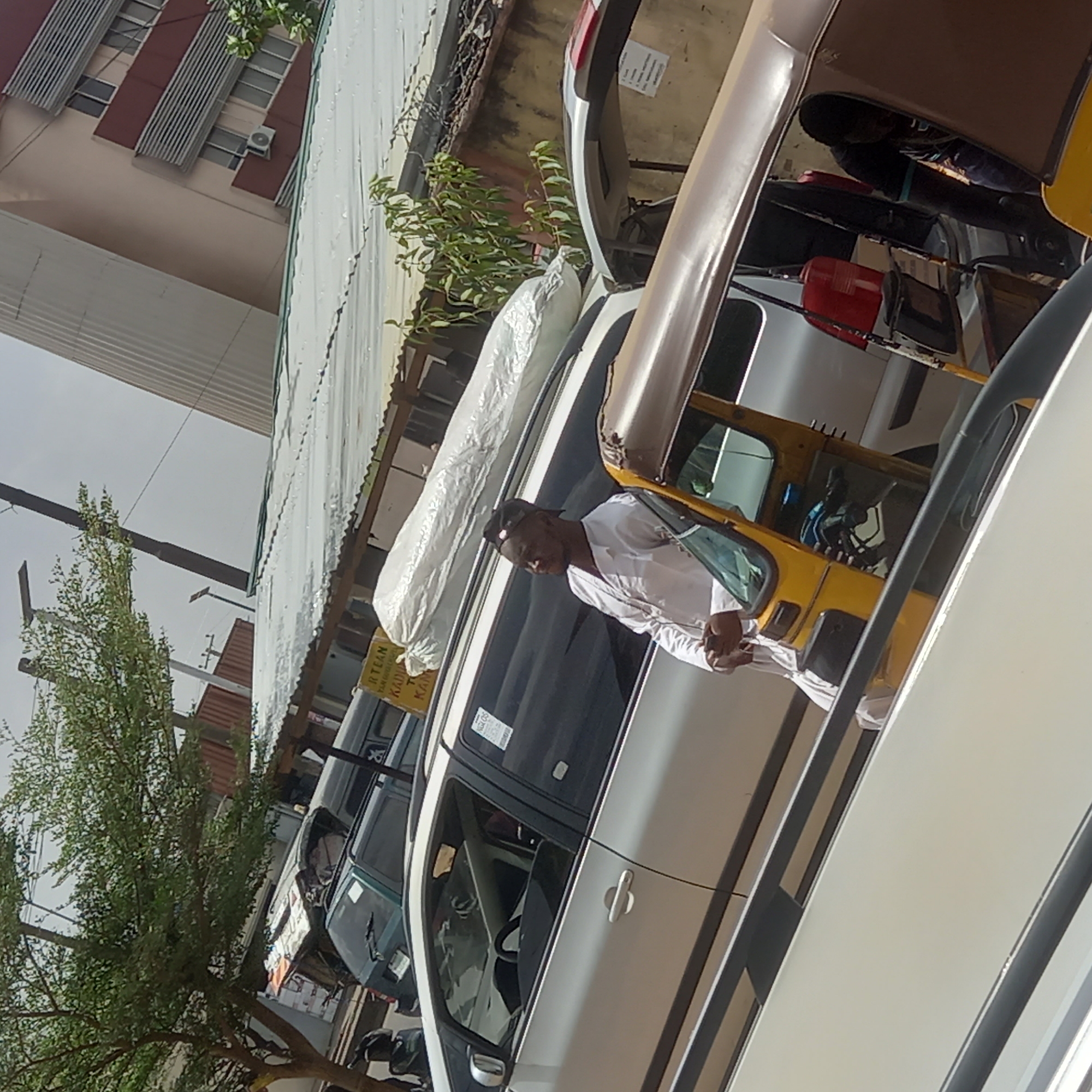 A daytime street scene in Dadi Riba, Nigeria, shows a man standing next to parked vehicles. The image is captured from a slightly elevated, rotated perspective.
The primary subject is a man of color, wearing a white long-sleeved shirt or traditional attire, with sunglasses resting on his head. He stands upright between a white car and a yellow tricycle (commonly known as a "keke napep" in Nigeria), looking directly at the camera with a neutral expression, his hands clasped in front of him.
The white car, likely an SUV or van, is parked with a large white, plastic-wrapped bundle secured on its roof rack. A yellow sign is tied to this rack, bearing the partially visible text "RTEAN", "YOUR COMFORT", and words starting with "KADI..." and "KANI...". A white sticker on the white car's side window shows the numbers "00105" along with other illegible text. Behind the man, the yellow tricycle is visible, with its interior partially obscured. In the background, other vehicles, including a dark green car, are parked.
The scene features a multi-story building with distinctive red/brown sections, silver louvers, and an air conditioning unit. Corrugated metal roofs are also visible. To the left, a large, leafy green tree adds natural elements to the urban setting. The sky is bright but appears overcast or hazy, suggesting either mid-day or an unclouded but diffused light condition. The overall impression is that of a bustling street or market area, common in Nigerian towns.